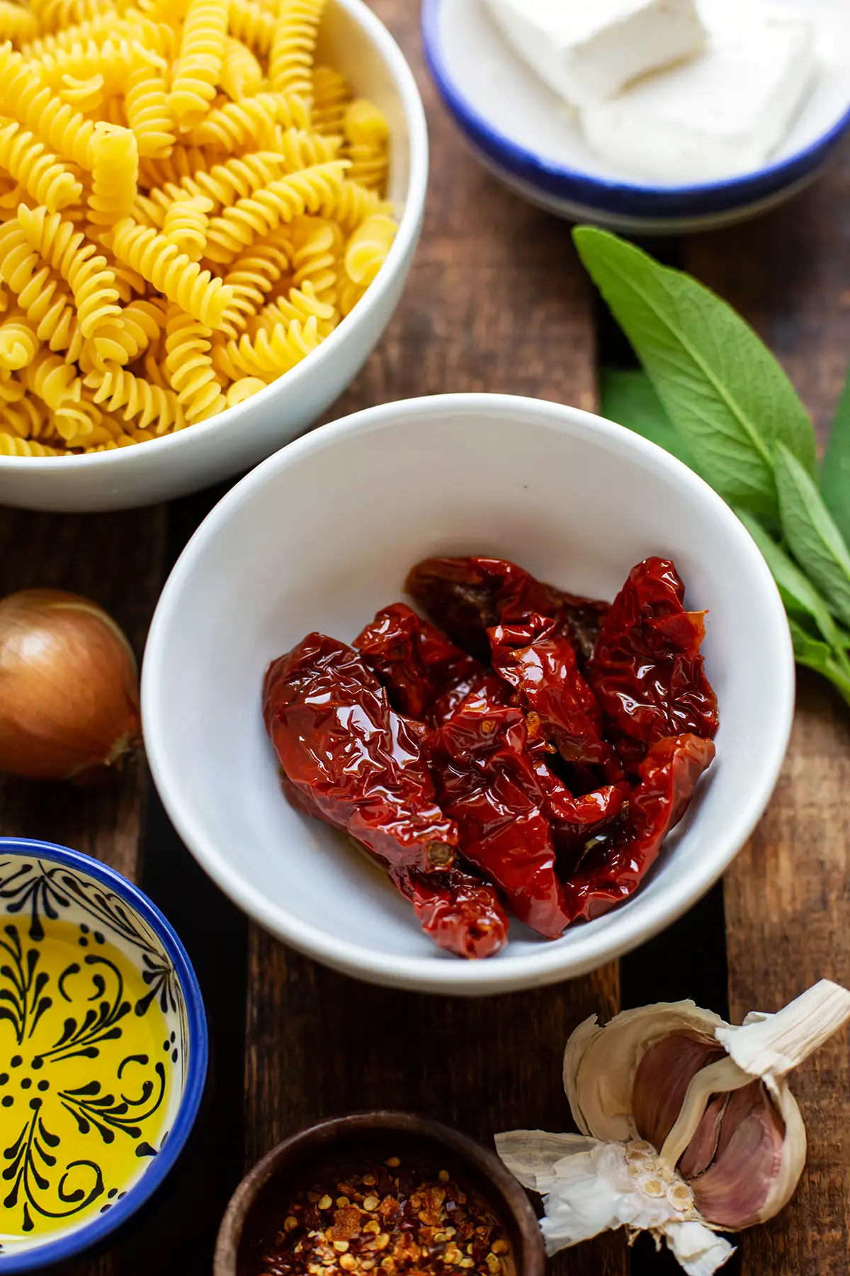 Zutaten für die italienische Salbei Pasta einzeln auf kleine Schüsseln oder lose aufgeteilt. Zu sehen sind getrocknete Tomaten, frischer Salbei, eine weiße Zwiebel, Fusilli, Öl, Knoblauchzehen, Chiliflocken und Feta.