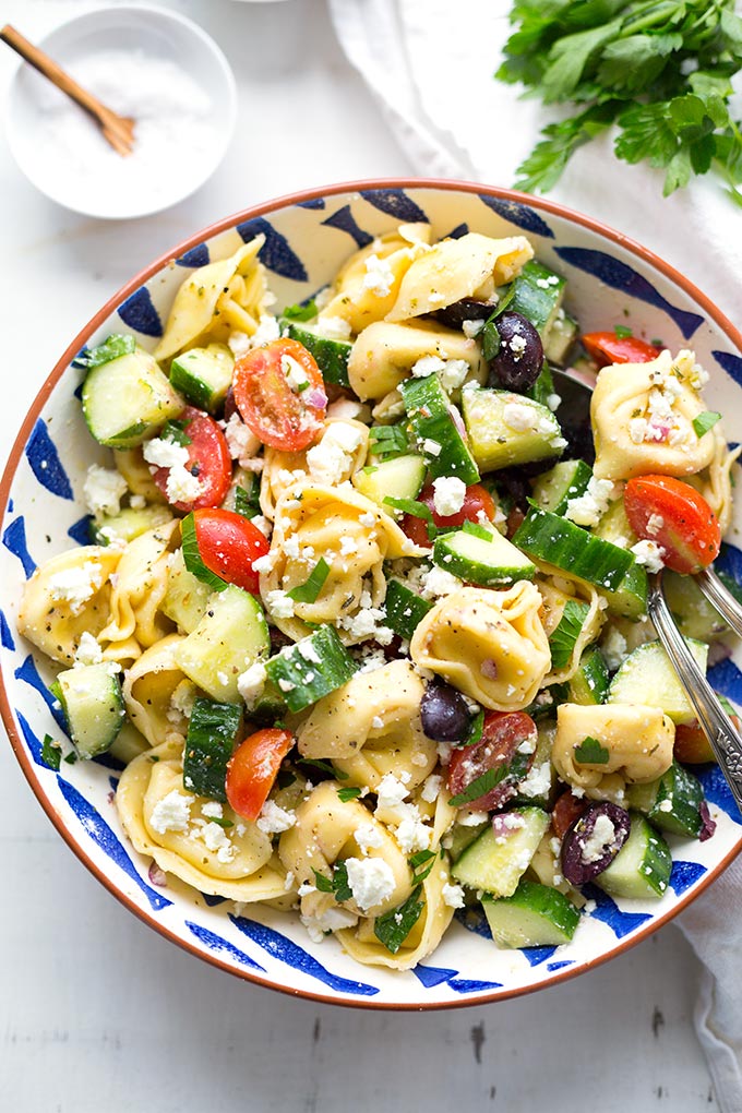 Griechischer Tortellinisalat in einer blau-weißen Schüssel mit Besteck. Die Schüssel steht auf einem hellen Untergrund. Zu sehen sind außerdem Petersilie und eine kleine weiße Schüssel.