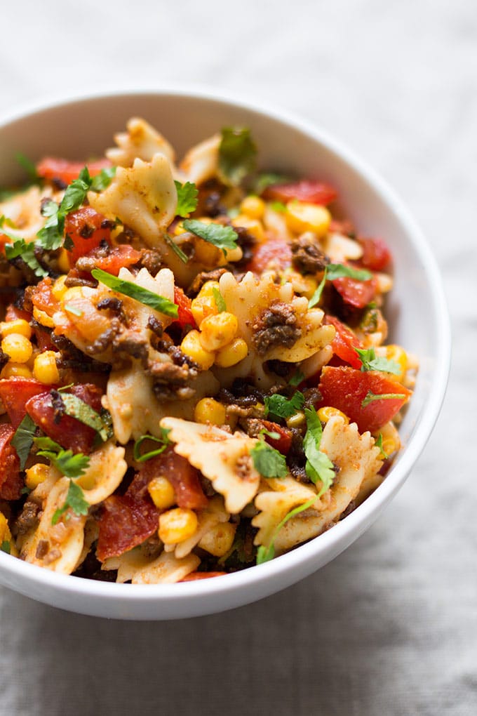 Einfacher Taco Nudelsalat mit würzigem Hackfleisch, Tomaten, Nudeln, Mais, Koriander und Taco Sauce in einer weißen Schüssel auf hellem Untergrund.