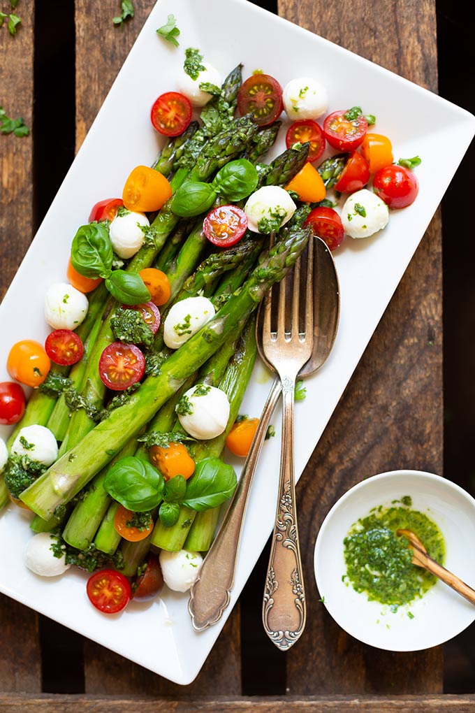 Fertiger Spargel-Caprese-Salat auf einem weißen Teller mit Gabel und Löffel auf dunklem Holzuntergrund. Garniert mit Basilikum. Daneben eine kleine weiße Schüssel mit grünem Pesto.