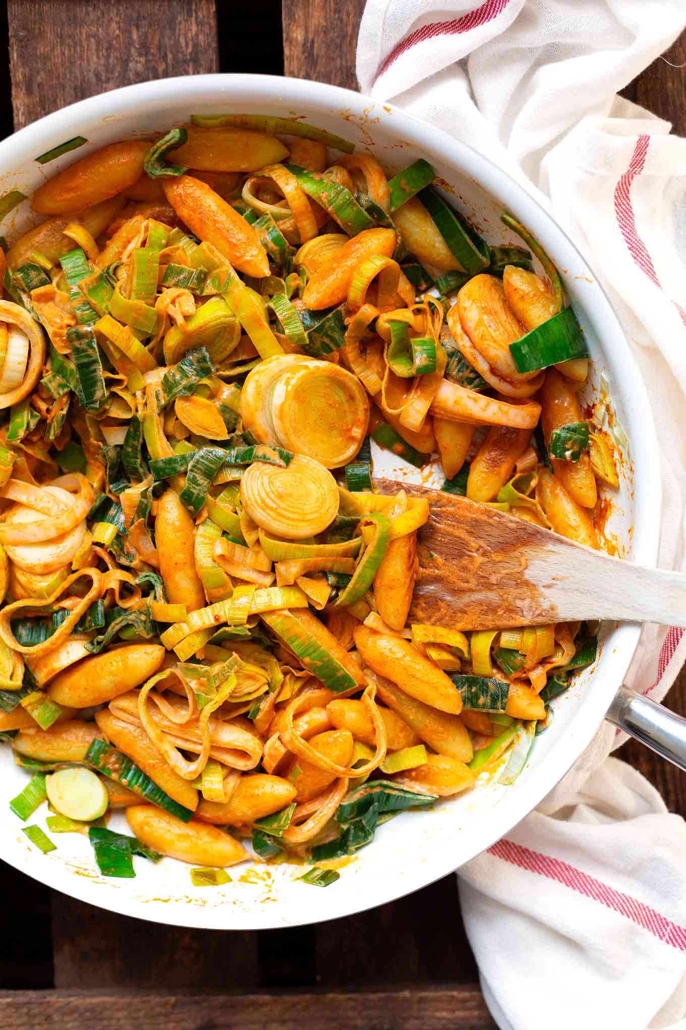 Fertige Lauch-Schupfnudel-Pfanne als One Pot Rezept in einer weißen Pfanne auf dunklem Holzuntergrund. Daneben ein weißes Geschirrtuch und ein Holzlöffel zum Umrühren in  der Pfanne.