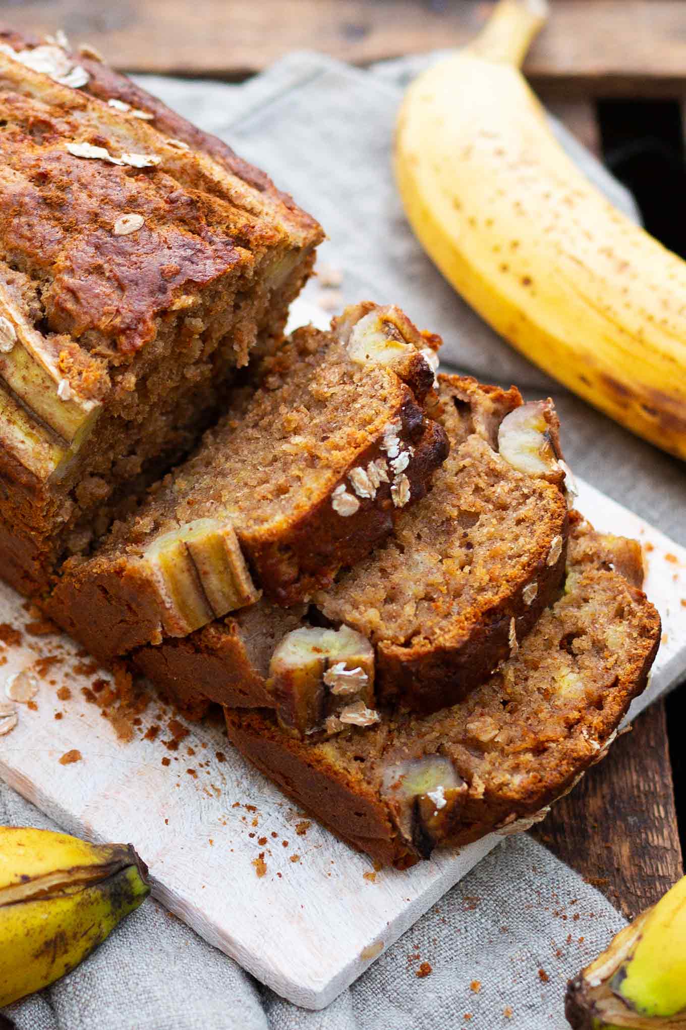 Aufgeschnittenes Bananenbrot ohne Zucker auf einem Holzbrett. Drumherum Bananen, Dinkelflocken und Zimt