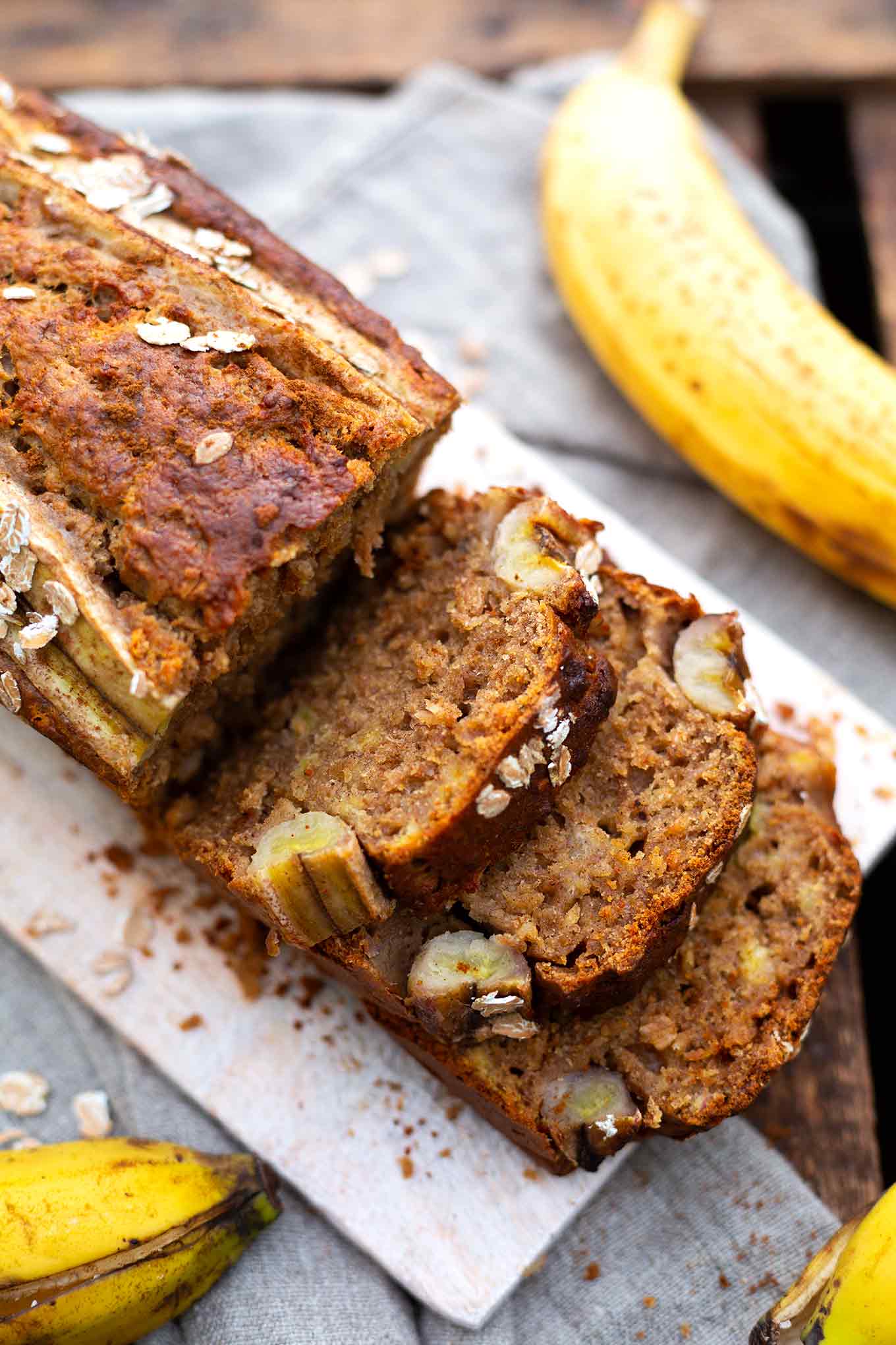 Aufgeschnittenes Bananenbrot ohne Zucker auf einem Holzbrett. Drumherum Bananen, Dinkelflocken und Zimt