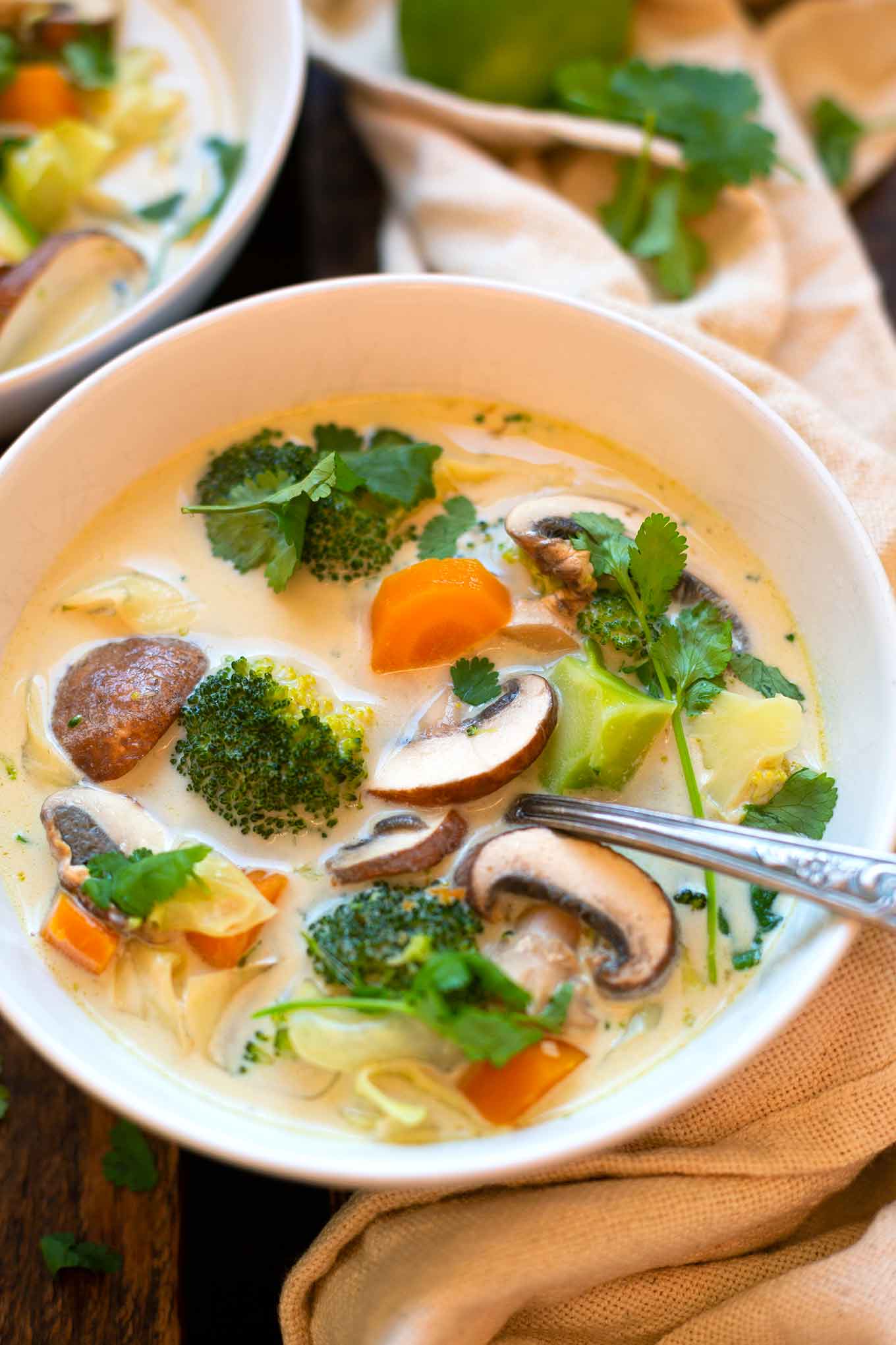 Thai-Kokos-Suppe mit Gemüse in einer weißen Schüssel. In der Suppe enthalten sind Brokkoli, Karotten, Pilze, Spitzkohl und obendrauf Koriander.