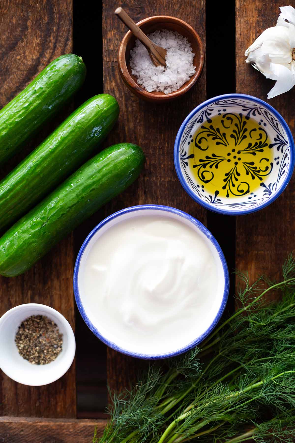Zutaten für Gurkensalat mit Joghurt-Dressing auf einem dunklen Holzuntergrund. Zu sehen sind, Öl, Gurken, Salz, Pfeffer, Knoblauch, Dill und Naturjoghurt.