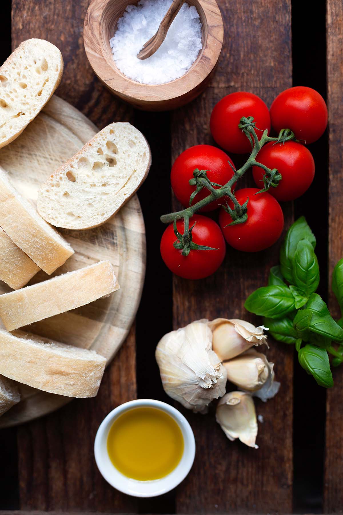 Alle Zutaten für das italienische Bruschetta auf einem dunklen Holzuntergrund. Man findet reife Tomaten, Knoblauch, Basilikum, Olivenöl und Ciabatta Brot