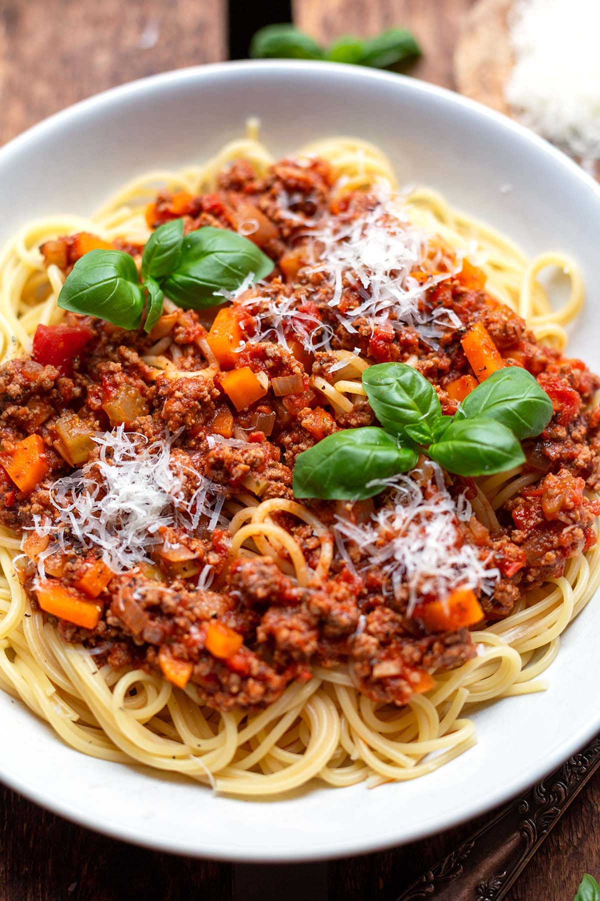 Spaghetti mit Bolognese Sauce angerichtet auf einem weißen Teller angerichtet mit Parmesan und Basilikum auf einem dunklen Holzuntergrund.