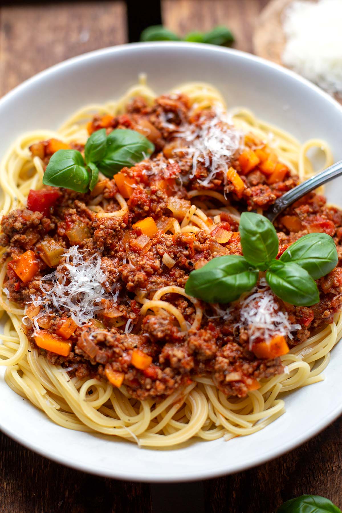 Spaghetti mit Bolognese Sauce angerichtet auf einem weißen Teller angerichtet mit Parmesan und Basilikum auf einem dunklen Holzuntergrund.