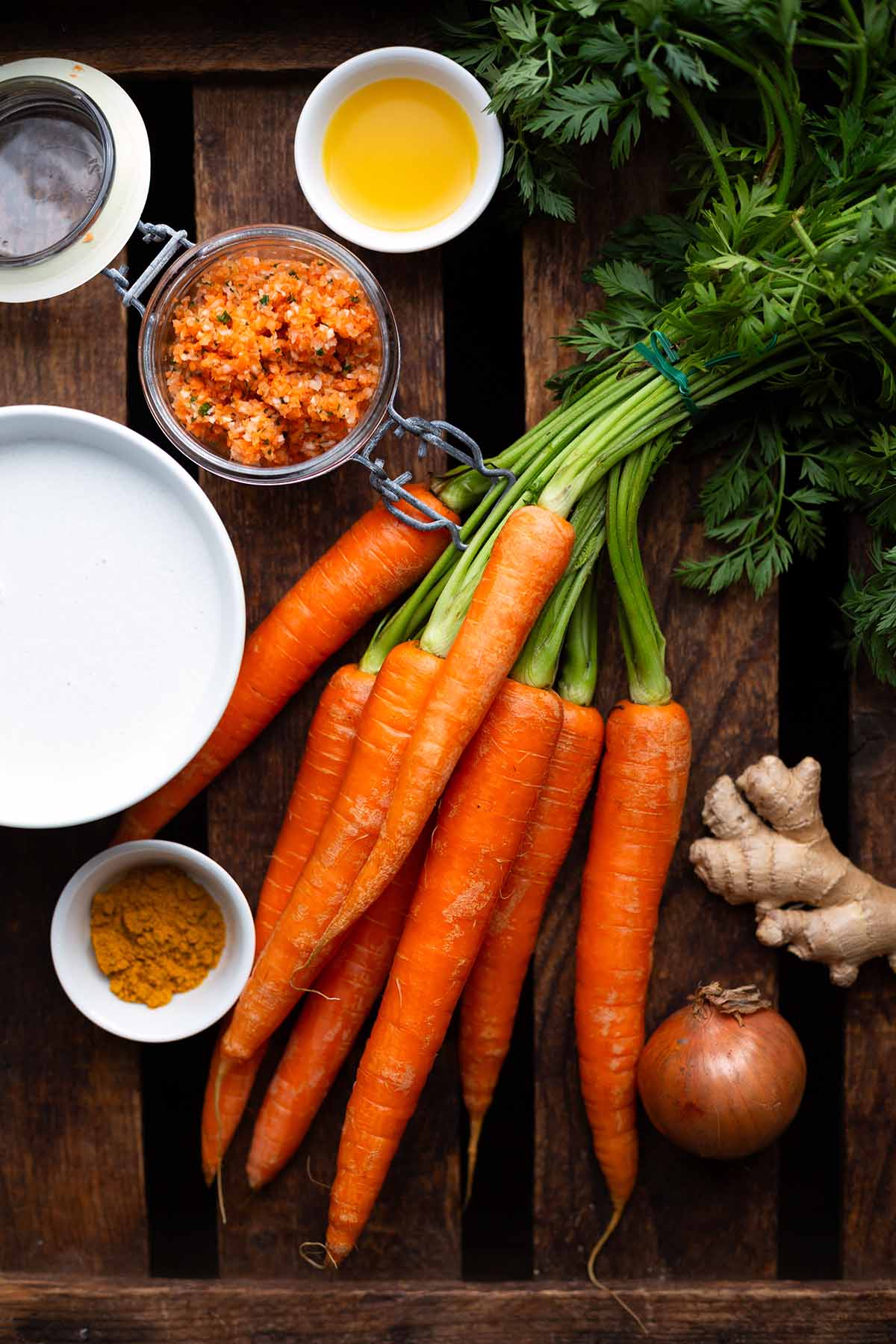 Die Zutaten für die vegane Möhrensuppe auf einem dunklen Holzuntergrund. Zu sehen sind Karotten, Gemüsebrühe, Kokosmilch in einer Schüssel, Ingwer, Currypulver, Öl in einer kleinen weißen Schüssel, eine Zwiebel.