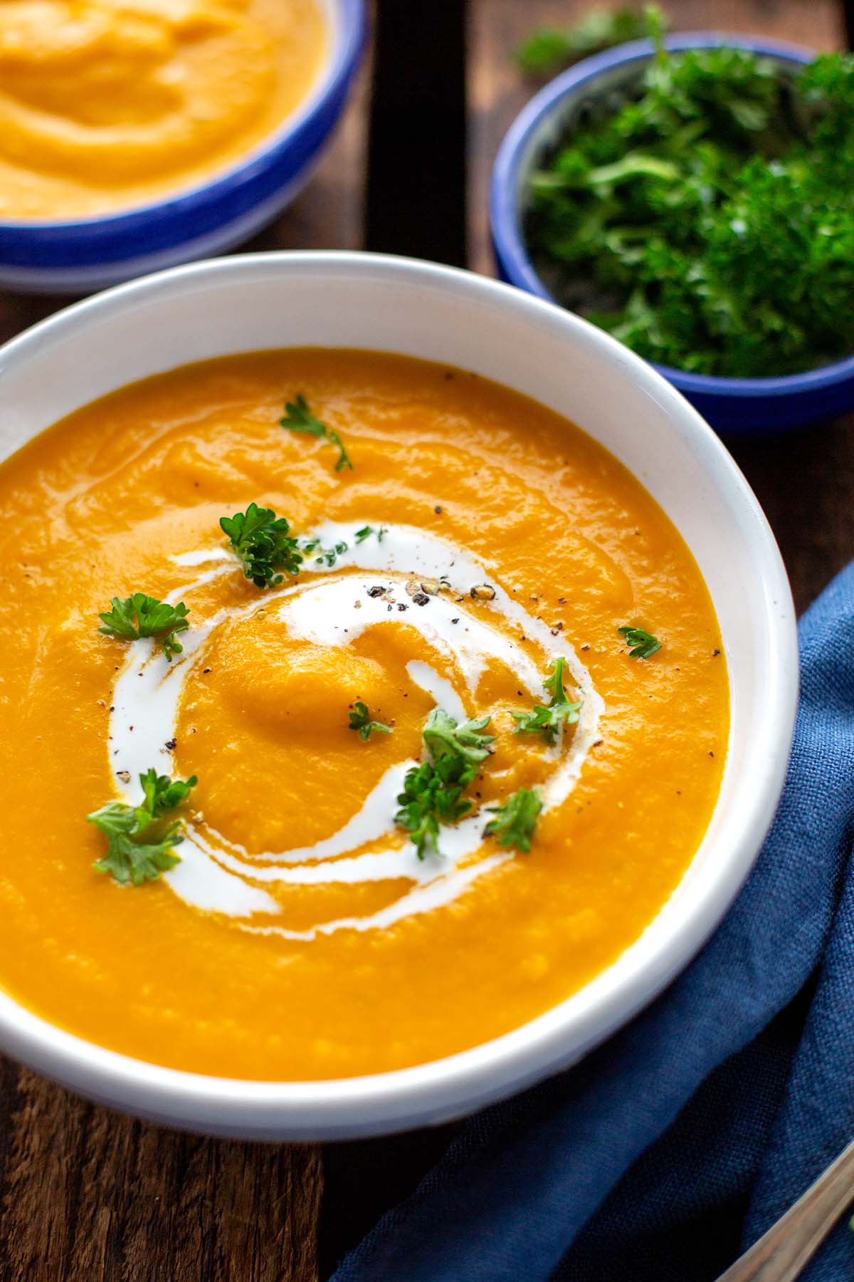 Fertige Möhrensuppe mit Ingwer und Kokosmilch in einer weißen Schüssel. Garniert mit Petersilie. Im Hintergrund ein blaues Geschirrtuch und weitere vegane Möhrensuppe und Petersilie einzeln in blauen Schüsseln.