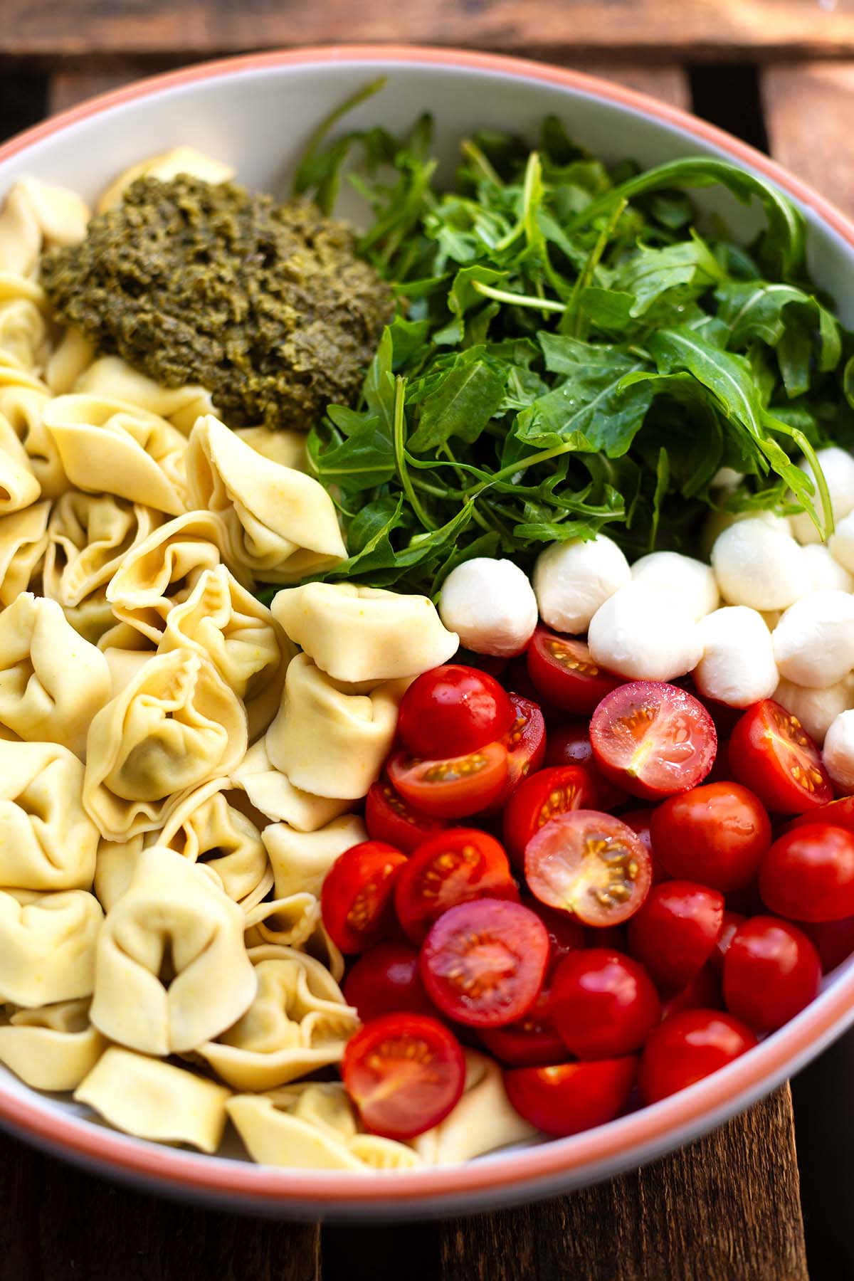 Alle Zutaten für das Tortellinisalat Rezept in einer großen Schüssel.
Zu sehen sind: Cherrytomaten, Rukola, Mozzarella-Kügelchen, grünes Pesto und Tortellini.