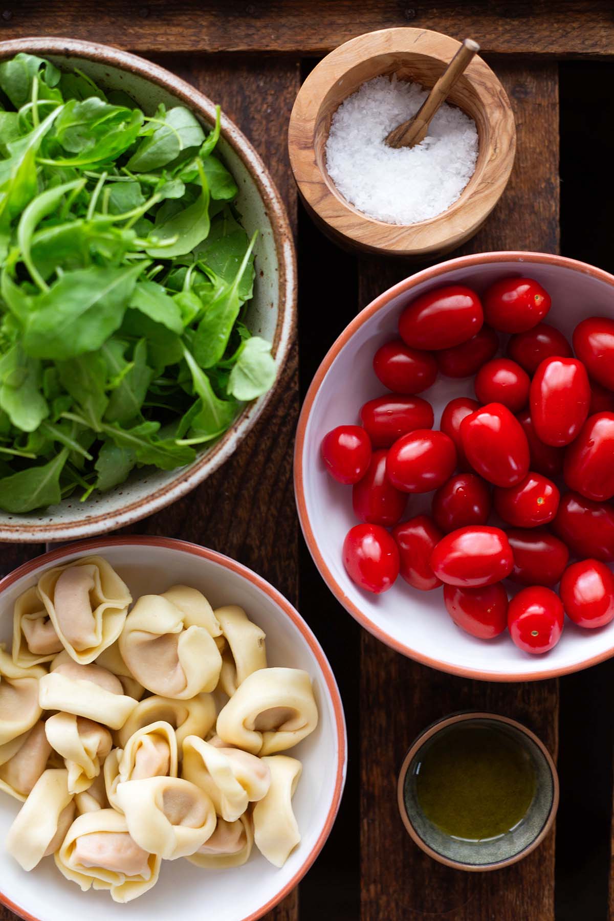 Zutaten für Tortellini-Pfanne auf einer Holzkiste: Tortellini, Rucola, Tomaten, Parmesan, Olivenöl und Salz
