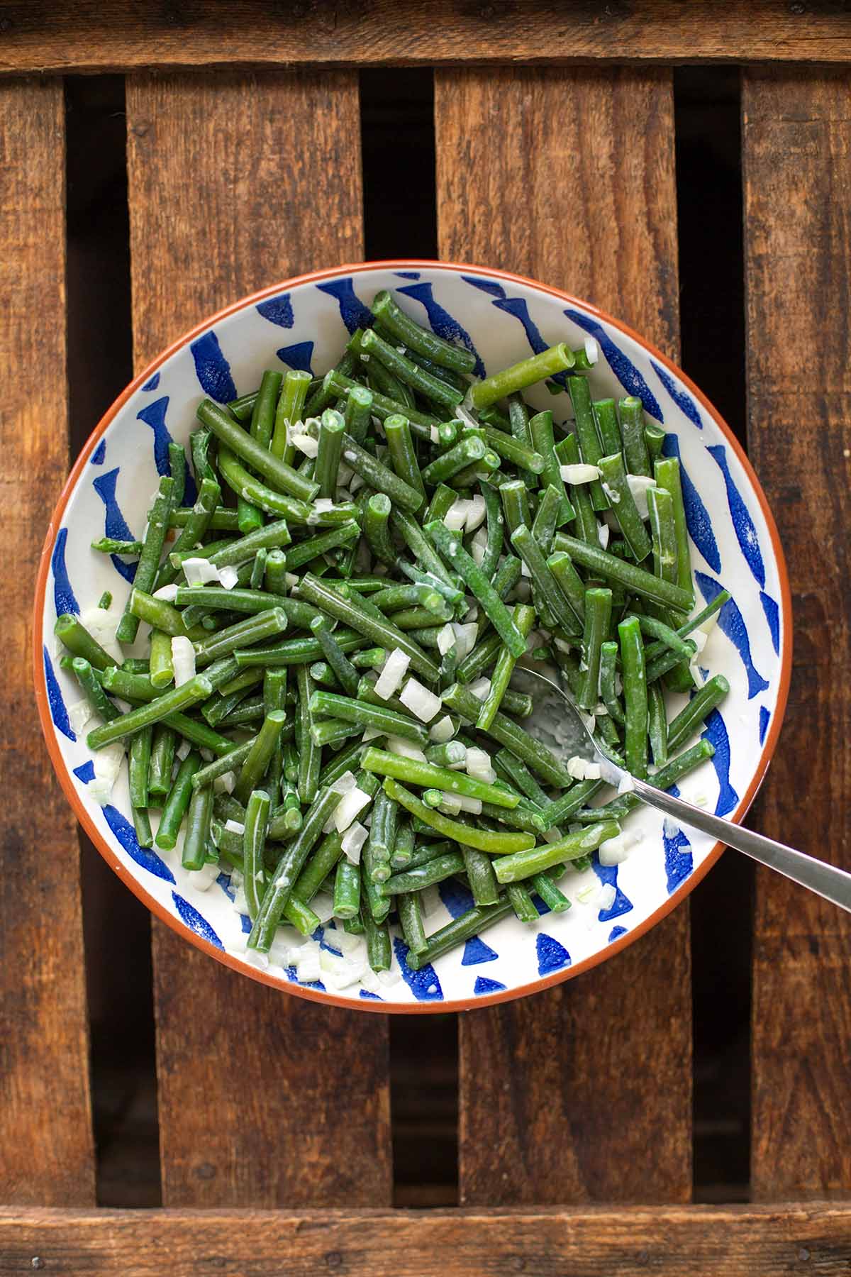 Zutaten für den Grüne Bohnen-Salat vermischt in einer weiß-blauen Schüssel auf dunklem Holzuntergrund. Ein Löffel liegt halb in der Schüssel.