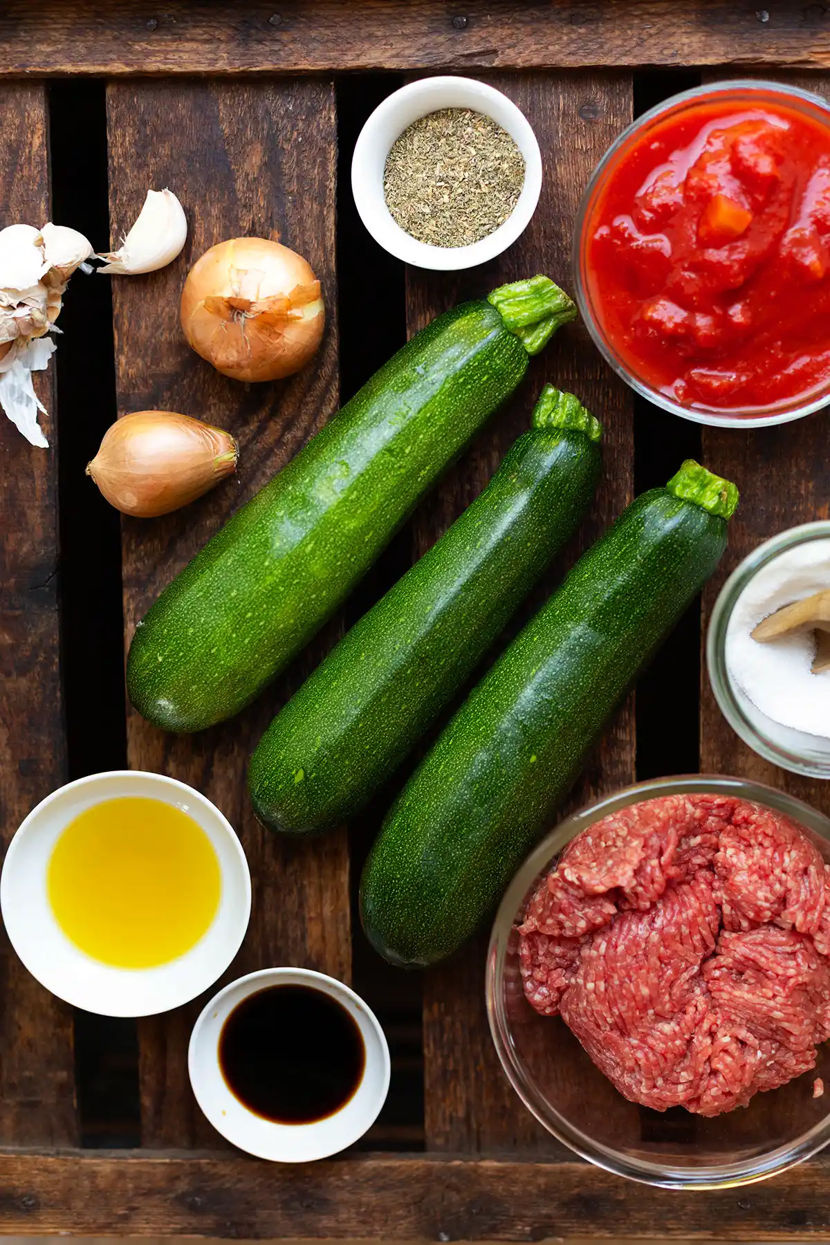 Ingredients for the zucchini minced meat pan in individual bowls or directly on a dark wooden surface. You can see three zucchini, chopped tomatoes, minced meat, onion, garlic, Ö, soy sauce and spices.