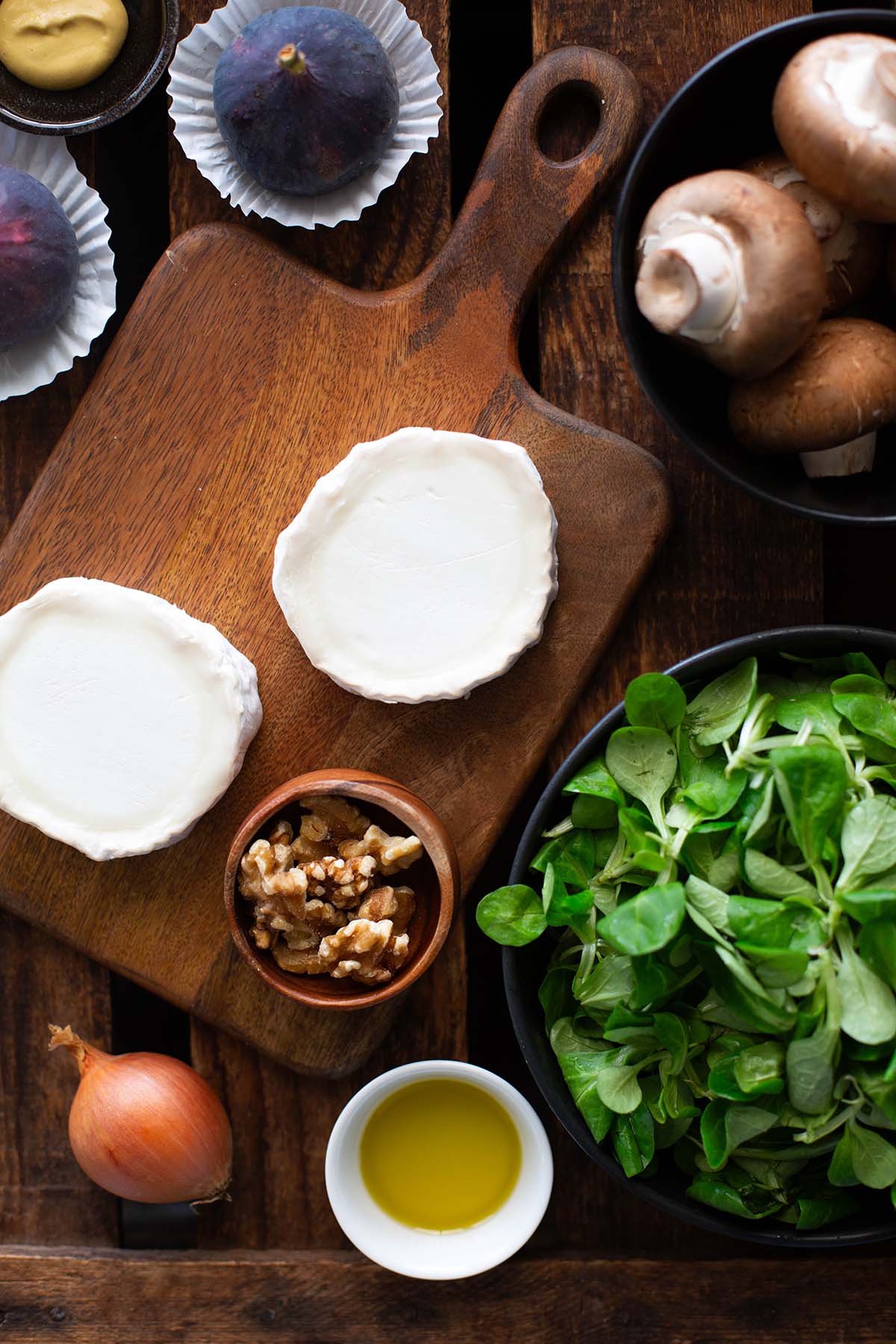 Zutaten für den Salat mit Ziegenkäse auf einem dunklen Holzuntergrund.
Zu sehen sind, Ziegenkäse, Feldsalat, Walnüsse, Senf, Feigen, Champignons, Zwiebel, Honig.