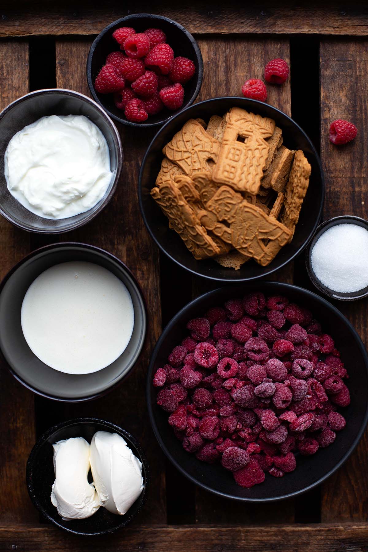 Zutaten für das Spekulatius-Rezept in einzelnen Schüsseln. Zu sehen sind Spekulatius-Kekse, Mascarpone, Joghurt, Sahne, Zucker, gefrorene Himbeeren, frische Himbeeren.