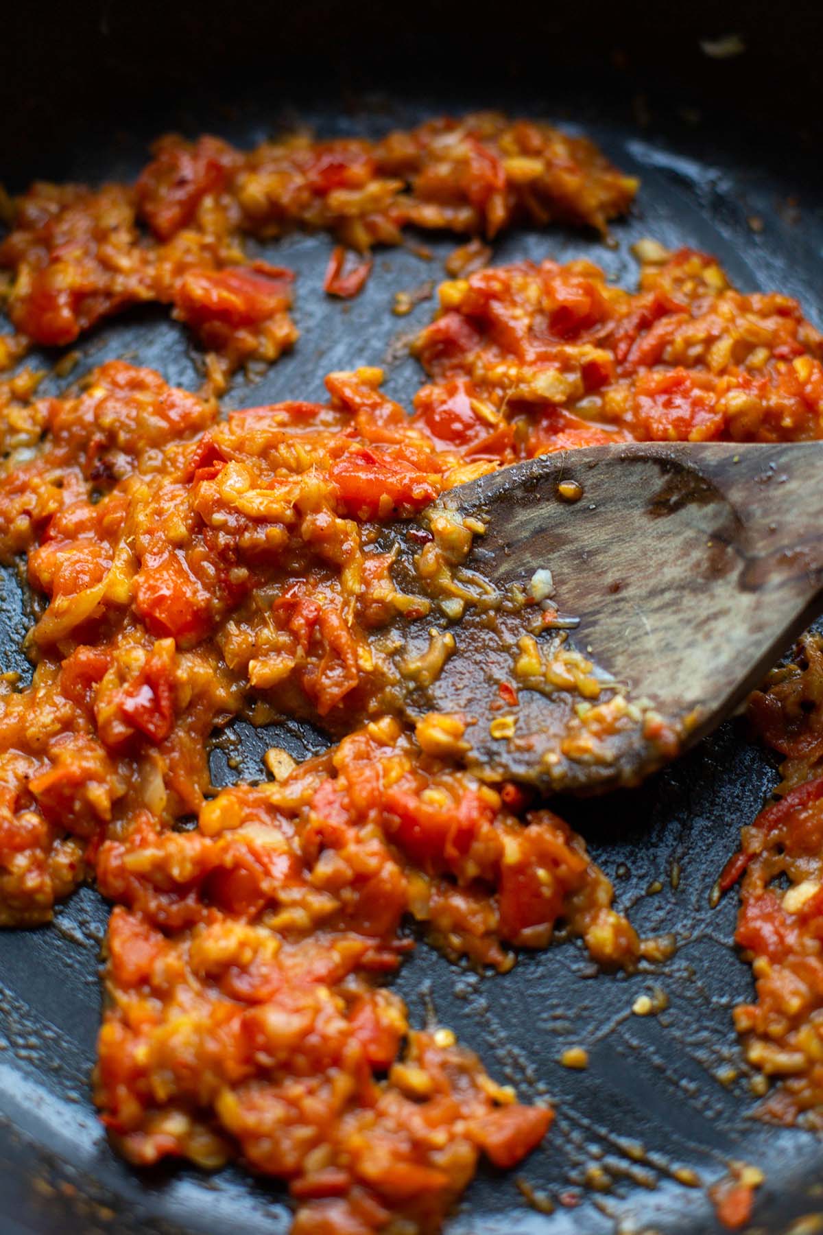 Tomaten, Zwiebel, Knoblauch und Ingwer in einer Pfanne, eingekocht mit Holzkochlöffel.