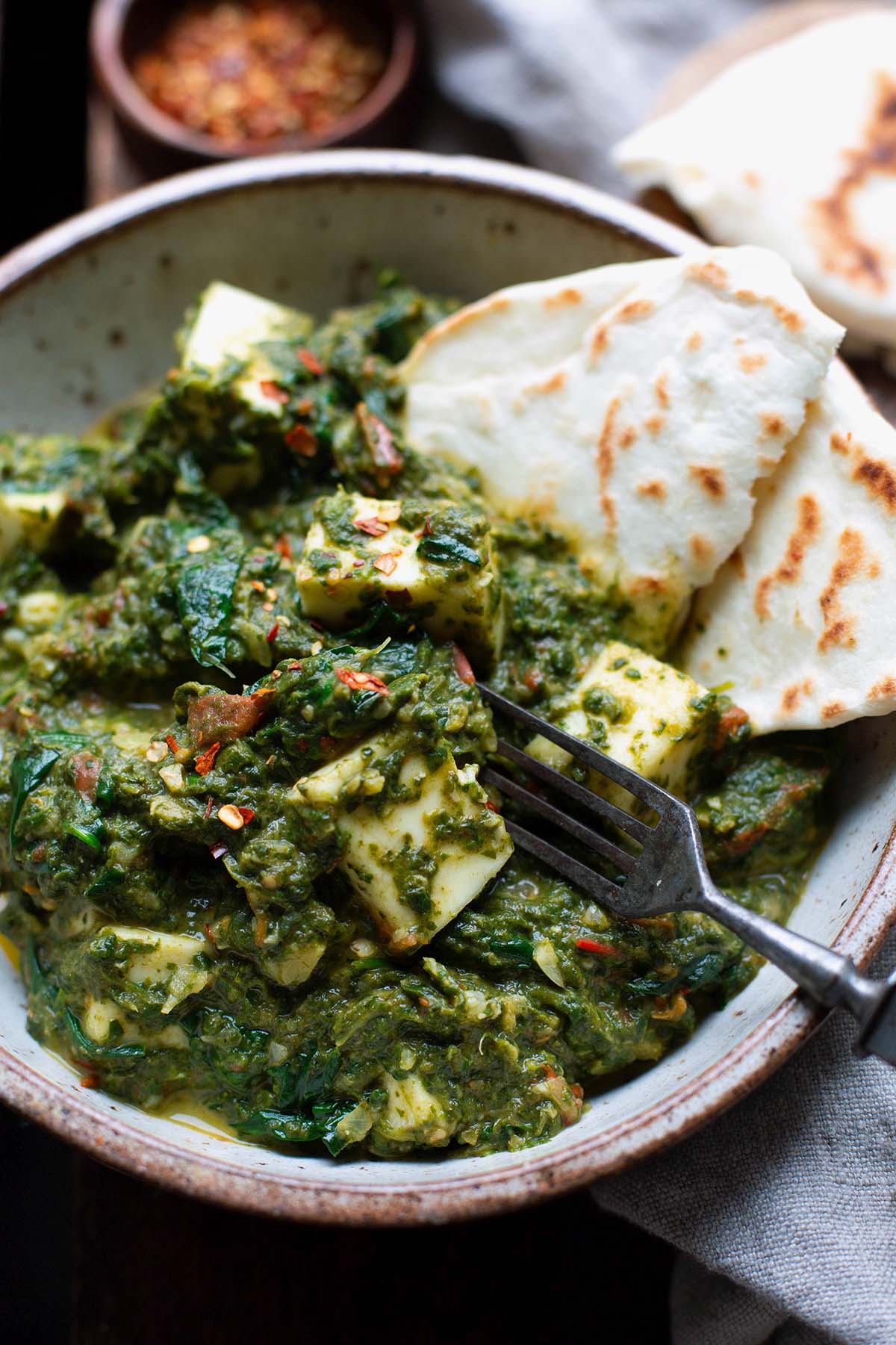 Indisches Palak Paneer mit Spinat und indischem Frischkäse in einer Schüssel. Dekoriert mit Chiliflocken und als Beilage Naan.