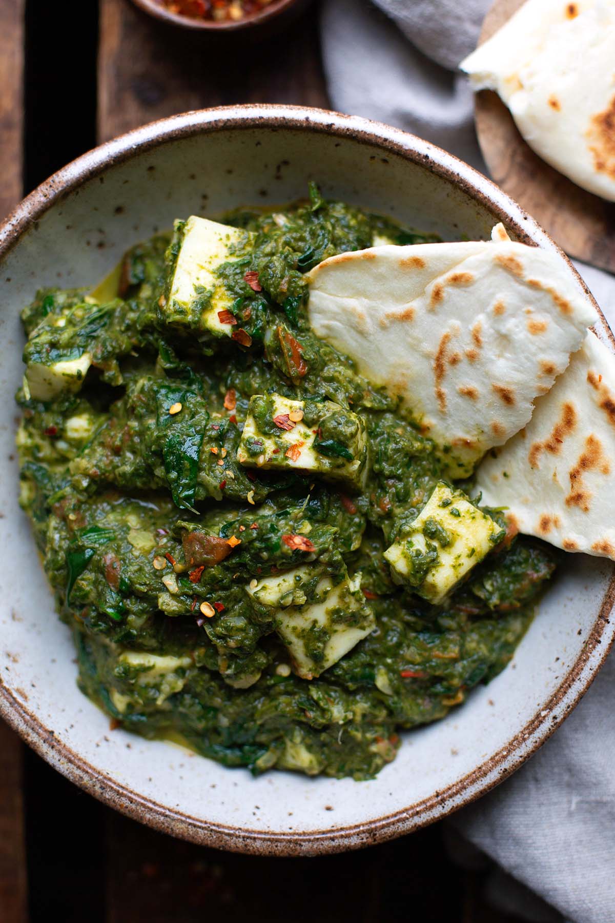 Palak Paneer mit Spinat und indischem Frischkäse in einer Schüssel. Dekoriert mit Chiliflocken und als Beilage Naan.