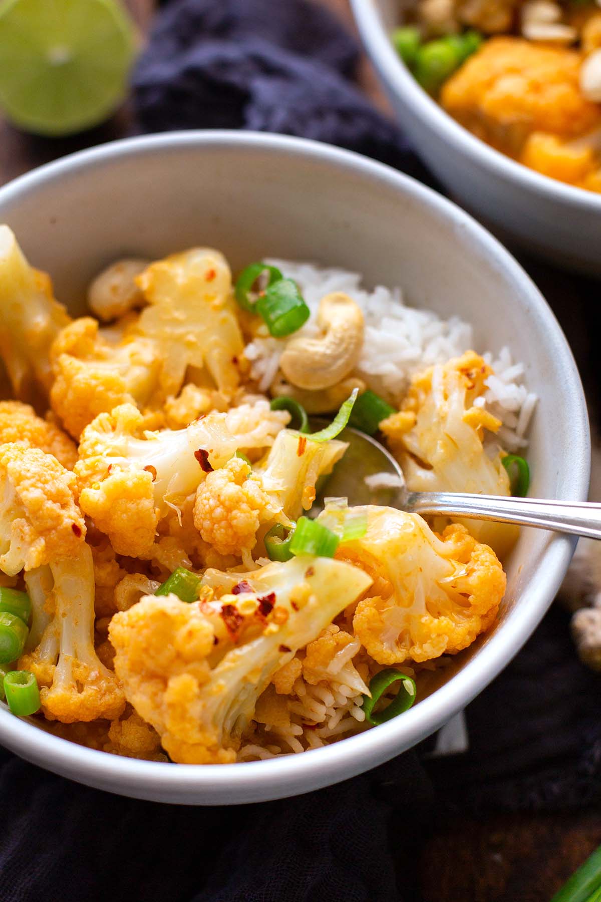 Fertiges veganes Curry in einer weißen Schüssel auf dunklem Holzuntergrund. Im Hintergrund ist noch eine zweite gefüllte Schüssel mit dem Blumenkohl-Curry zu sehen.