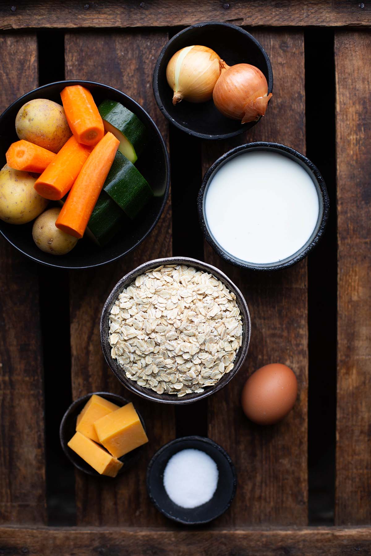 Zutaten für die Gemüsefrikadellen verteilt auf einzelne Schüsseln. Zu sehen sind Kartoffeln, Möhren, Zucchini, Haferflocken, Zwiebeln, ein Ei, Käse, Salz und Milch.