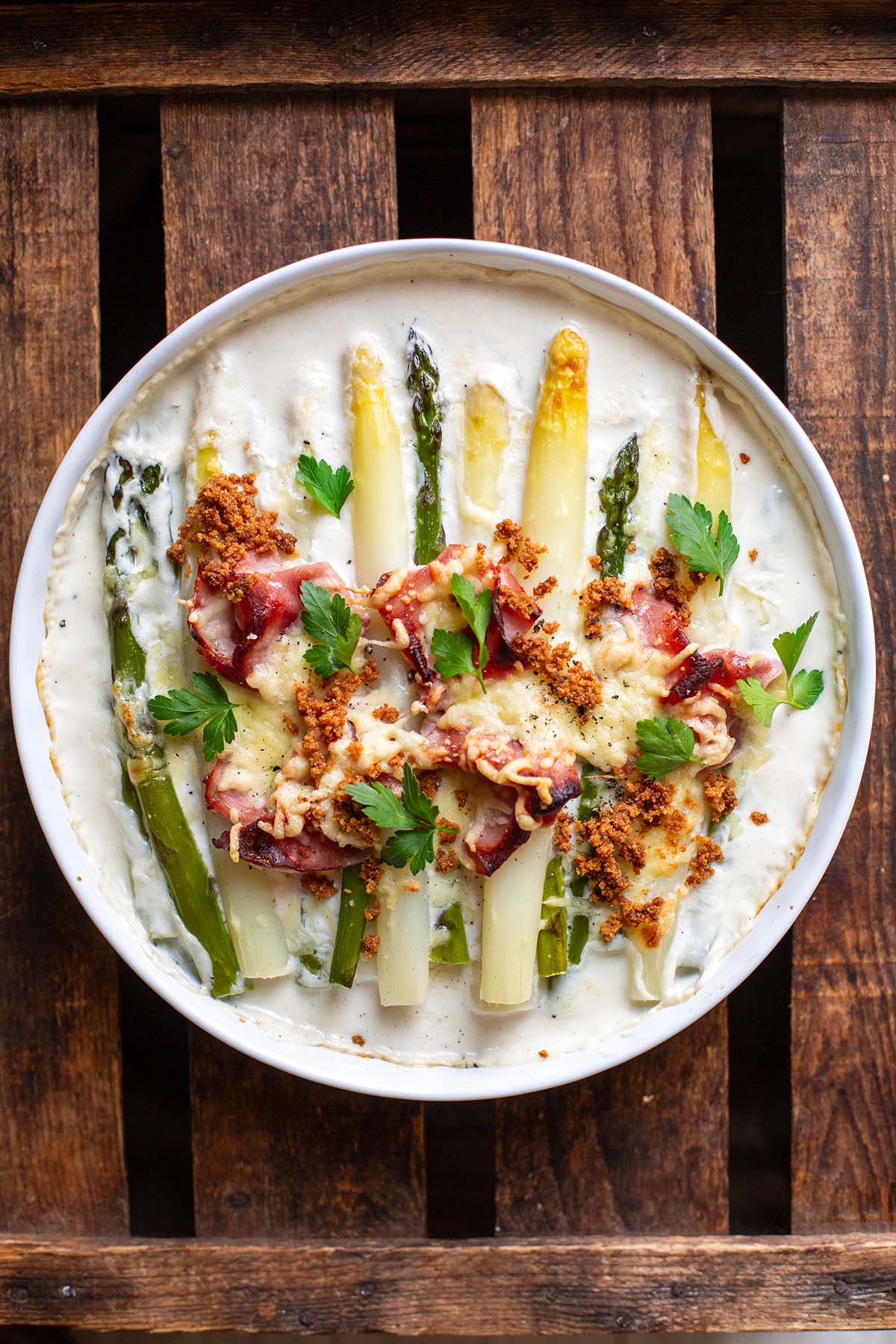 Überbackener Spargel mit Käse und Schinken auf einem weißen Teller auf dunklem Holzuntergrund.