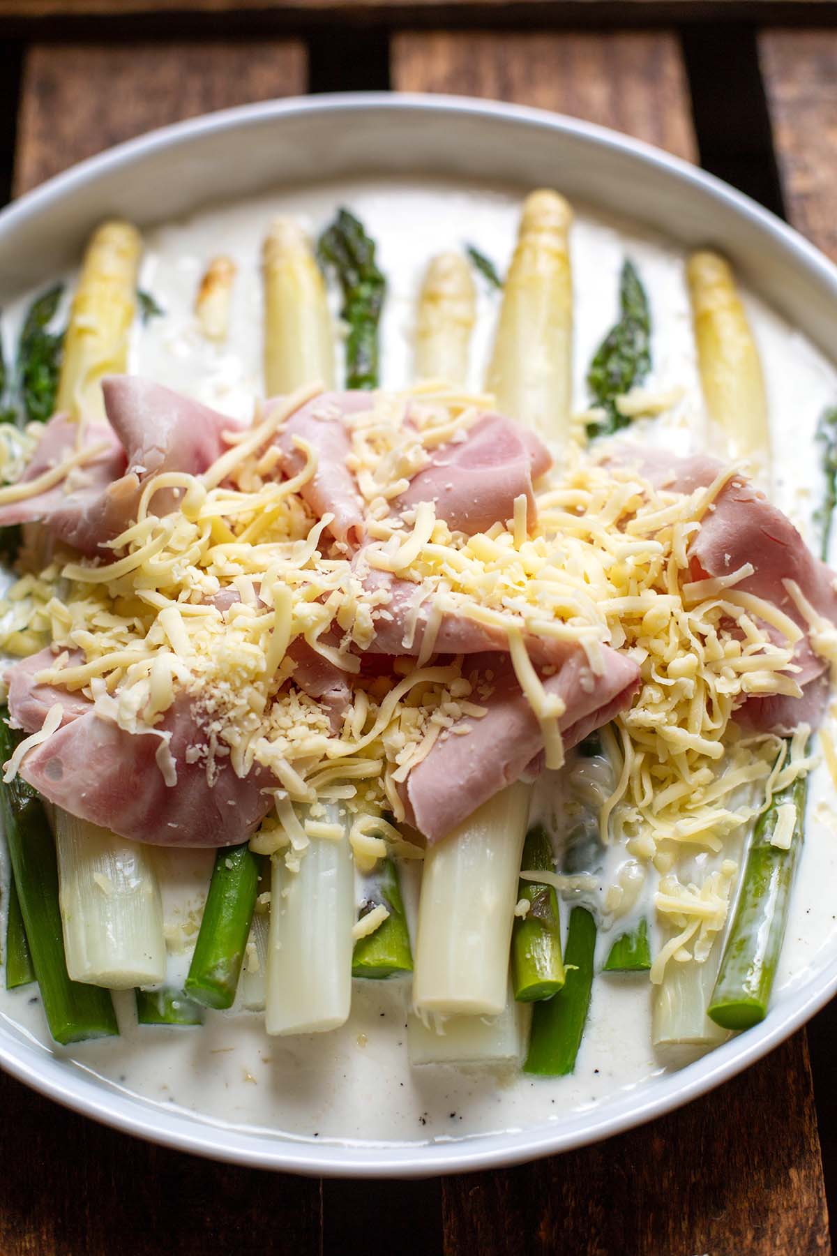 Grüner und weißer Spargel auf einem weißen Teller zusammen mit  Schinken, Käse und Béchamelsoße bevor das Ganze in den Backofen kommt.