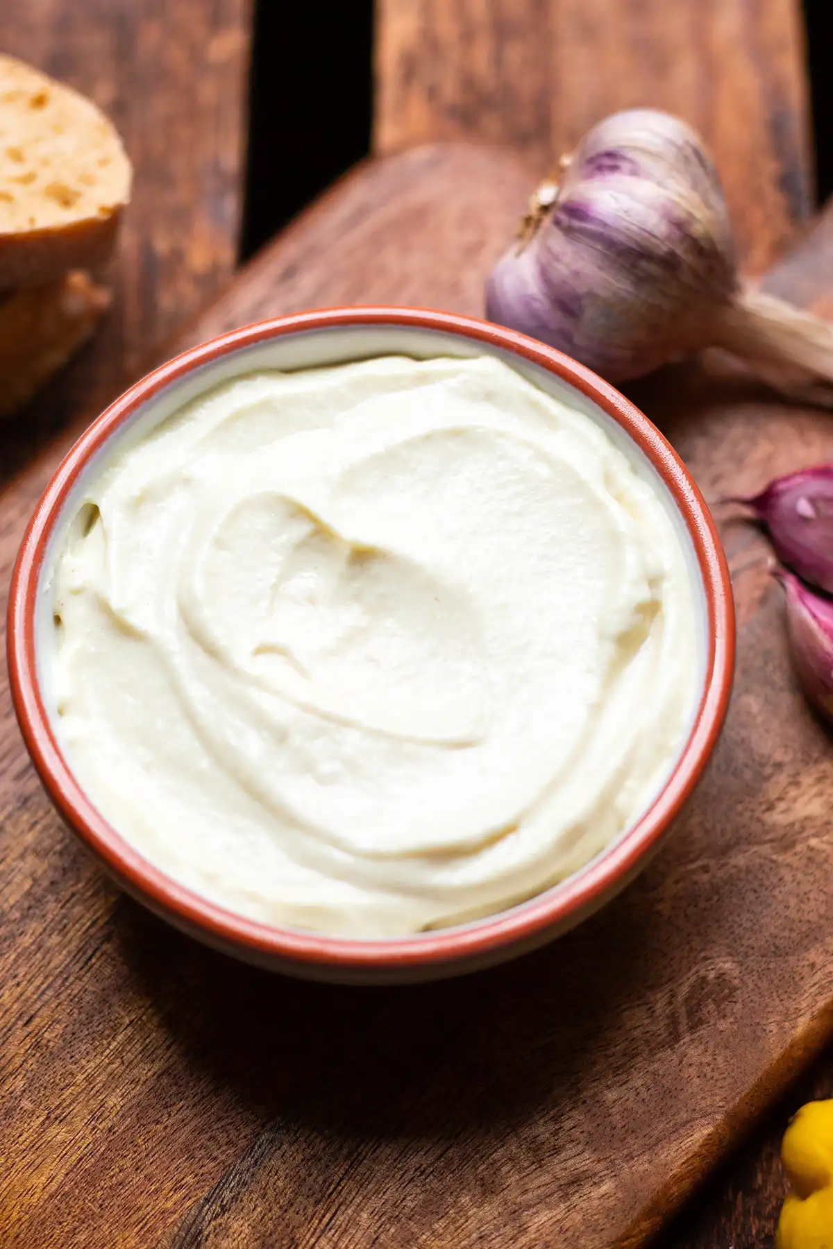Fertige Aioli in einer weißen Schüssel mit einem orangen Rand. Die Schüssel steht auf einem Holzbrett, welches ebenfalls auf einem dunklen Holzuntergrund steht. Im Hintergrund zu sehen ist Knoblauch, eine Zitrone und zwei Scheiben Baguettebrot.