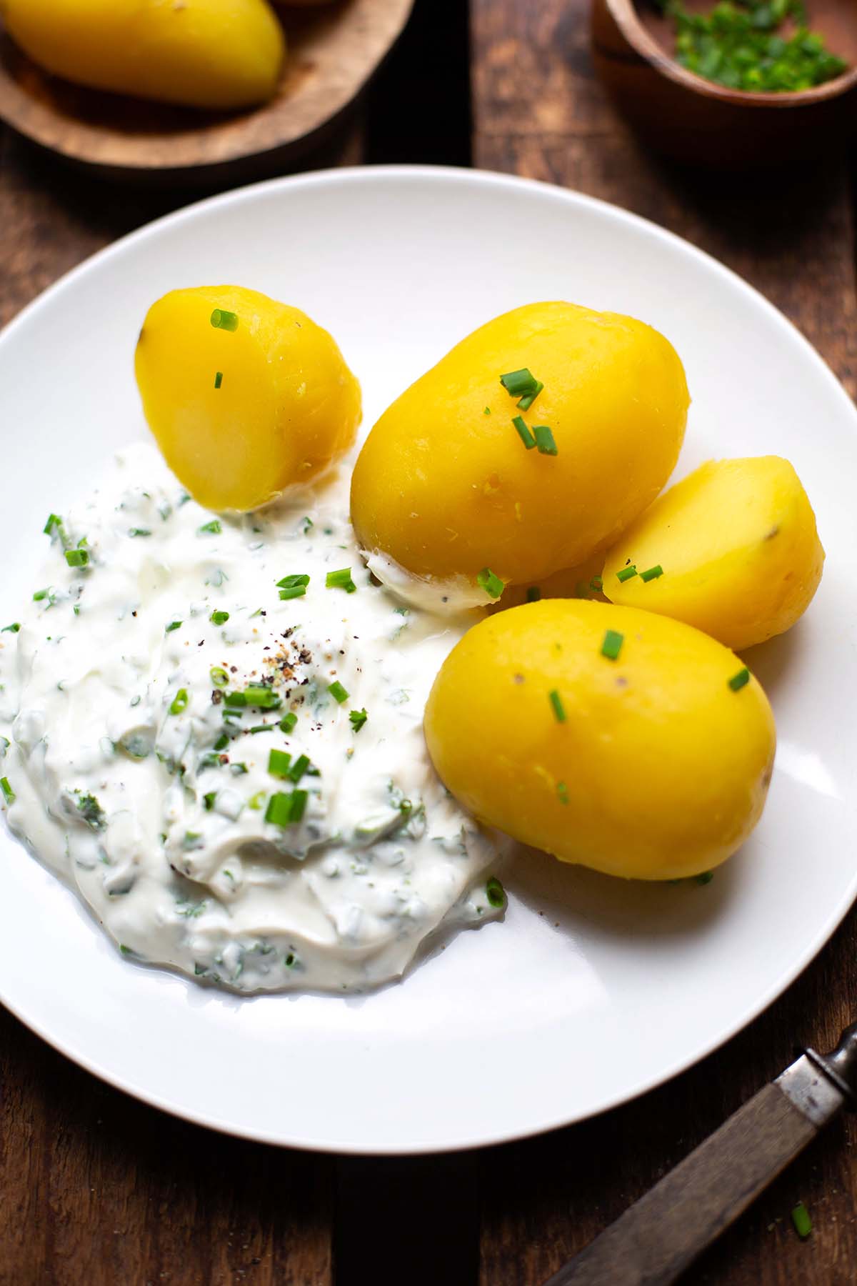 Fertige Kartoffeln mit Quark auf einem weißen Teller. Der Teller steht auf einem dunklen Holzuntergrund und im Hintergrund stehen noch zwei kleine Holzschälchen mit einmal Kartoffeln und einmal gehacktem Schnittlauch.