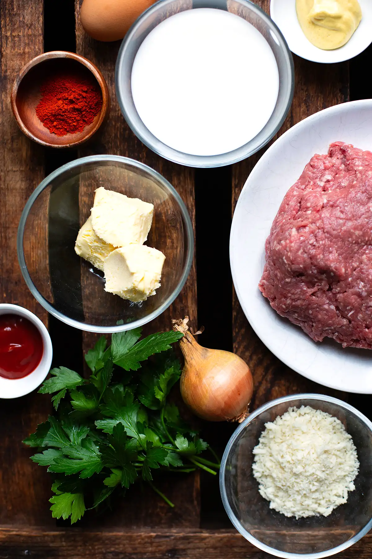 Zutaten für Omas Frikadellen Rezept in einzelnen Schälchen auf dunklem Holzuntergrund. Zu sehen sind gemischtes Bio-Hackfleisch, Milch, Butter, Ei, Ketchup, Senf, Paprikapulver, Petersilie und Panko-Paniermehl.