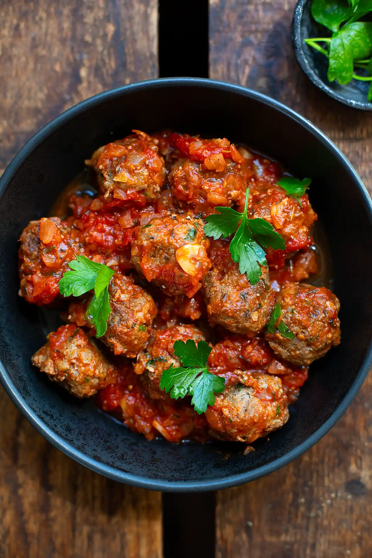 Fertige spanische Albondigas in einer schwarzen Schüssel. Diese steht auf einem dunkeln Holzuntergrund. Die Hackbällchen mit Tomatensauce sind mit glatter Petersilie garniert. Im Hintergrund steht ein zweites kleines Schälchen mit Petersilie.