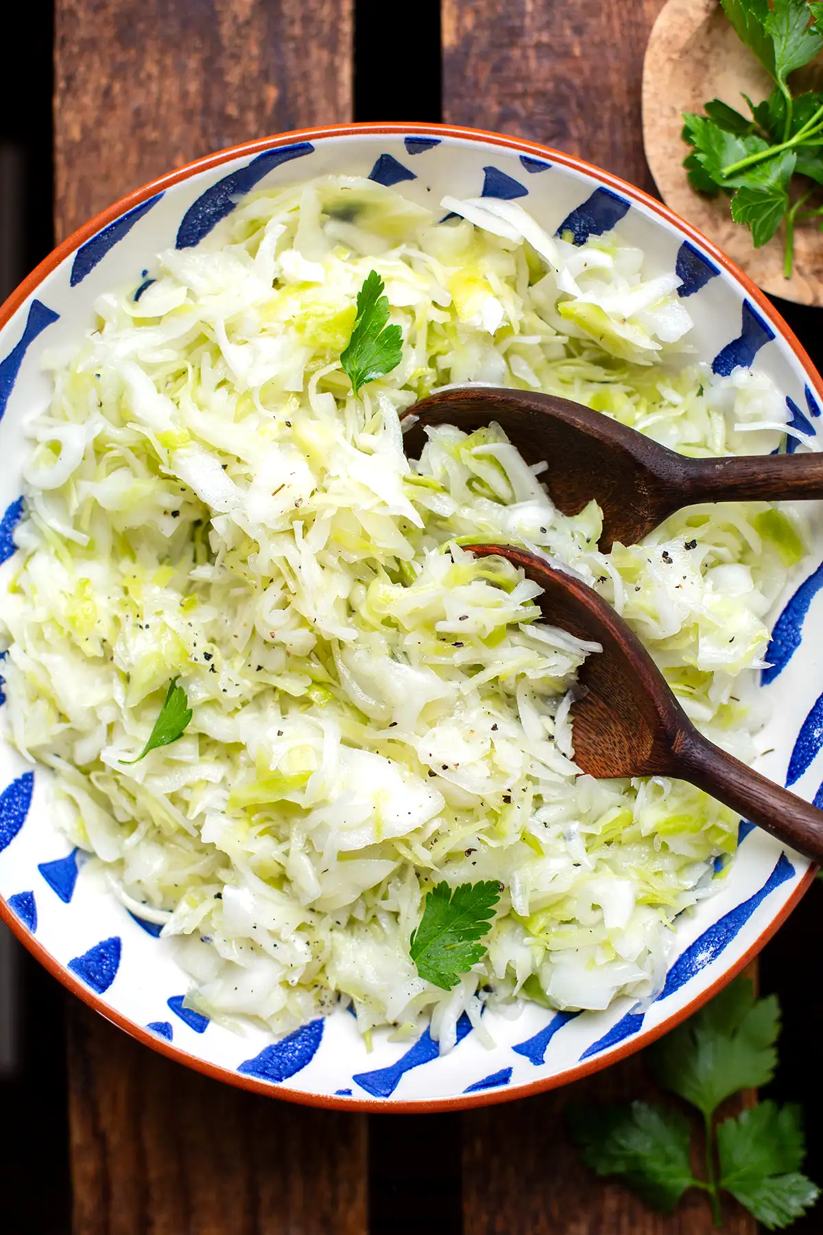 Fertiger griechischer Krautsalat in einer blau-weißen Schüssel mit braun-orangem Rand. Im Krautsalat stecken zwei große Holzlöffel zum Servieren. Die Schüssel steht auf einem dunklen Holzuntergrund. Im Hintergrund liegen ein paar Blätter glatte Petersilie.