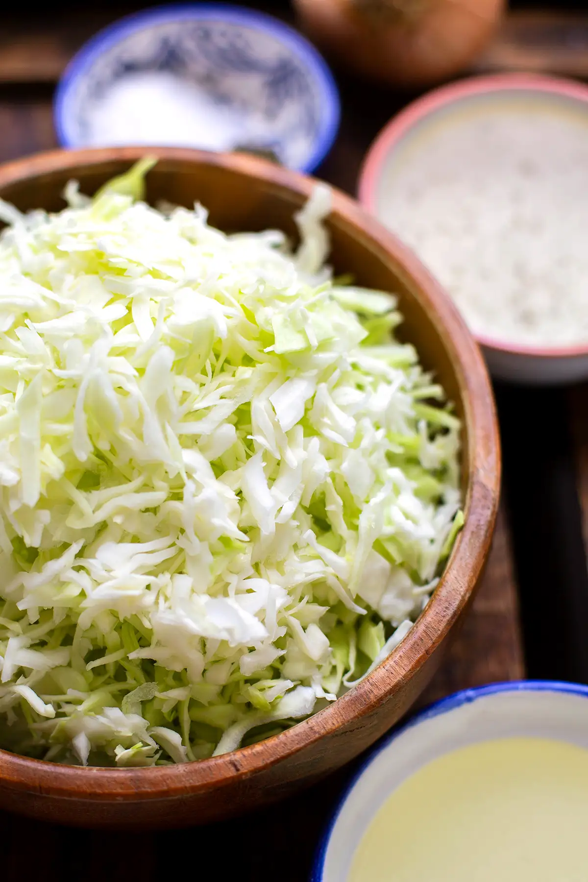 Zutaten für das Krautsalat Rezept einzeln in Schüsselchen auf dunklem Holzuntergrund. Zu sehen sind: gehobelter Weißkohl, eine weiße Zwiebel, Kräuteressig, Zucker.