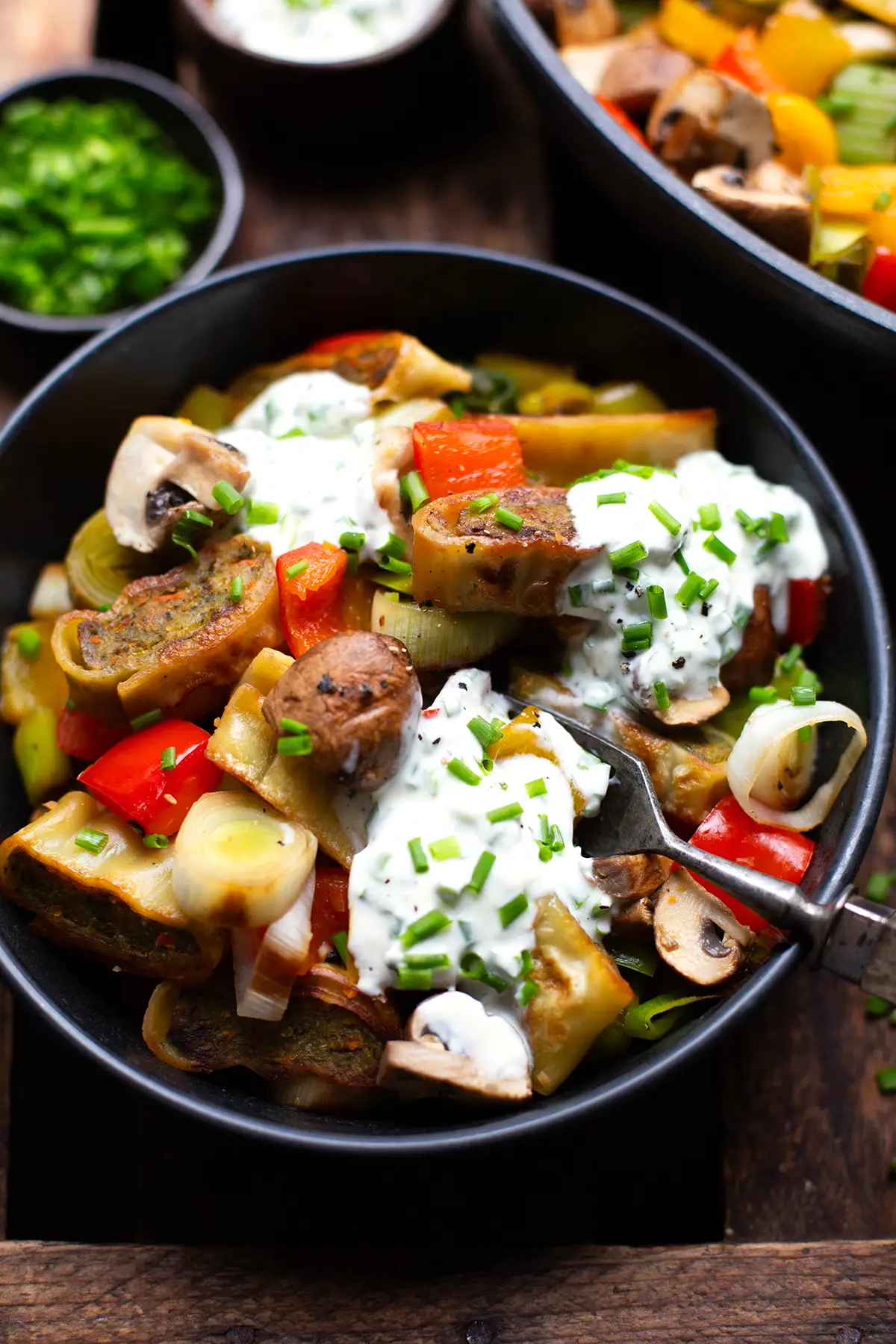 Fertige Maultaschen Pfanne mit Gemüse in einer schwarzen Schüssel. Garniert mit Schmand-Schnittlauch-Dip und einer Gabel. Im Hintergrund eine zweite gefüllte Schüssel und ein kleines Schälchen mit Schnittlauch. Die Schüsseln stehen auf einem dunklen Holzuntergrund.