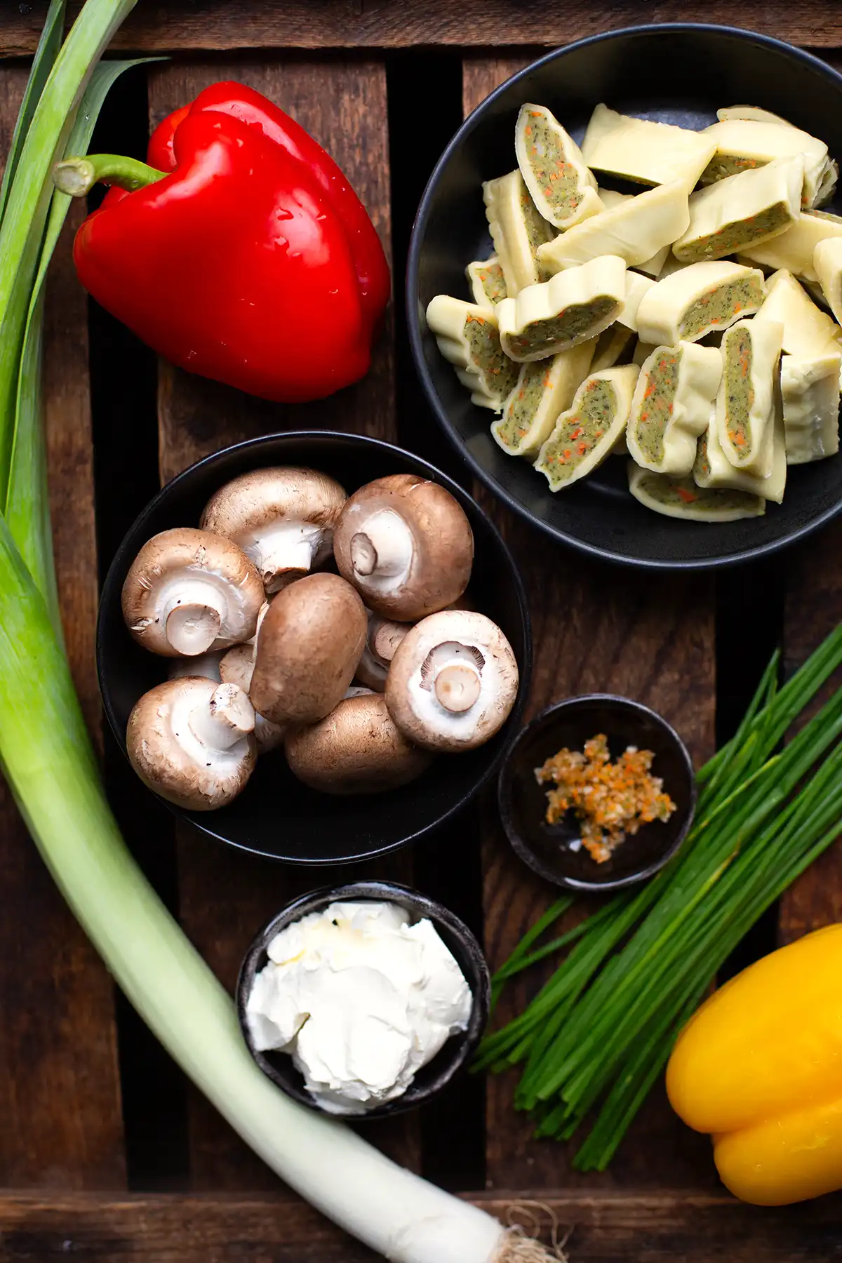 Zutaten für die vegetarische Maultaschen Pfanne mit Gemüse. Zu sehen sind: In Streifen geschnittene Maultaschen, eine rote Paprika, Lauch, braune Champignons, Gemüsebrühe-Paste, Schmand, eine gelbe Paprika und Schnittlauch.