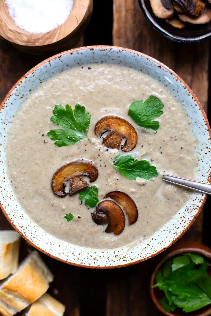 Fertige Pilzsuppe wie von Oma in einer Schüssel. Garniert mit glatter Petersilie und knusprig angebratenen Champignonscheiben. Daneben stehen zwei kleine Schüsselchen mit Salz und angebratenen Champignonscheiben.