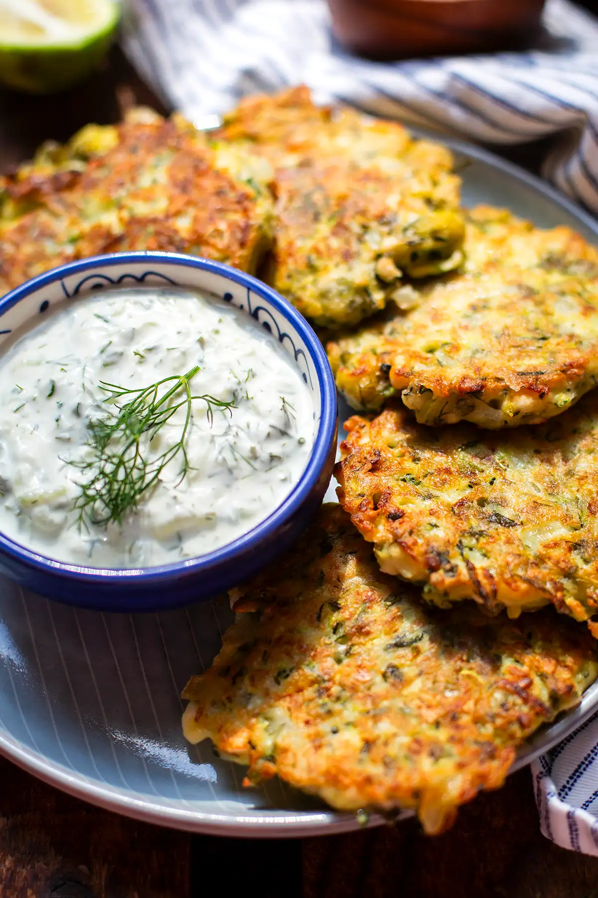 Fertige griechische Zucchinipuffer mit Feta auf einem Teller. Dazu eine kleine blau-weiße Schüssel garniert mit Dill.