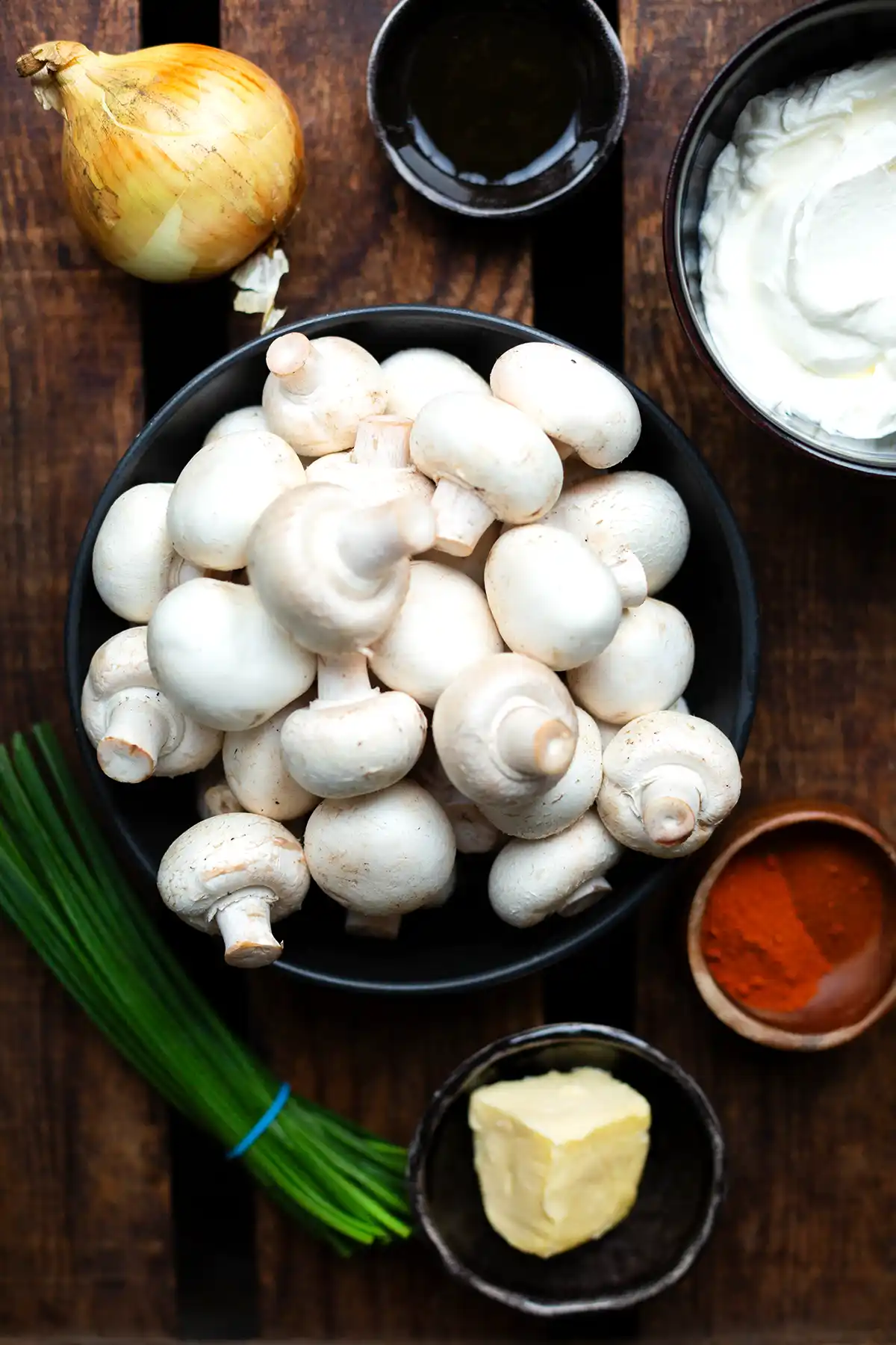 Die Zutaten für gebratene Champignons auf einen Blick. Zu sehen sind weiße Pilze, Schmand, Paprikapulver, Schnittlauch, Butter, Zwiebel und Ahornsirup.