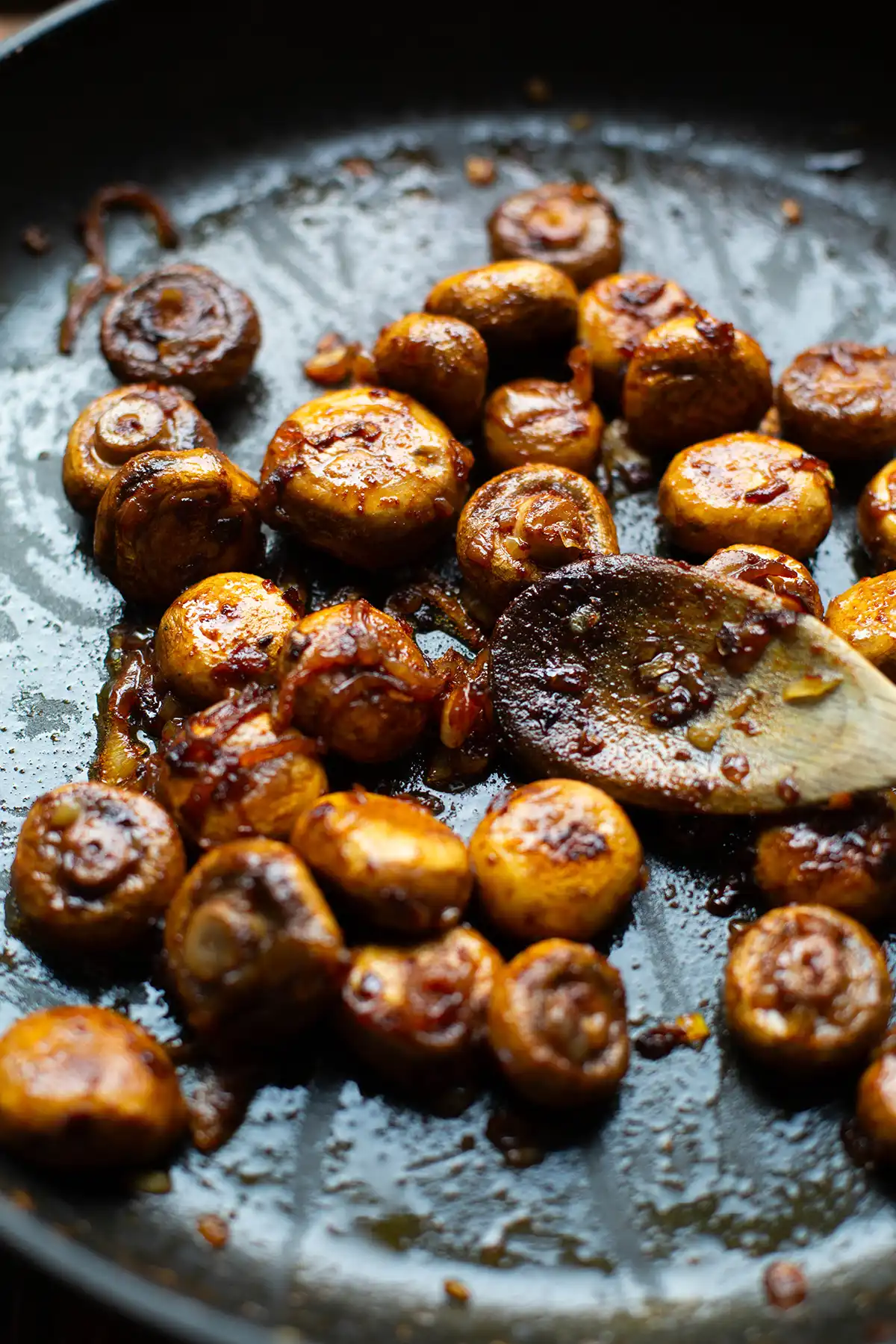 Champignonpfanne in der Zubereitung. Angebraten mit Zwiebeln und Butter in einer schwarzen Schmorpfanne mit Holzlöffel.