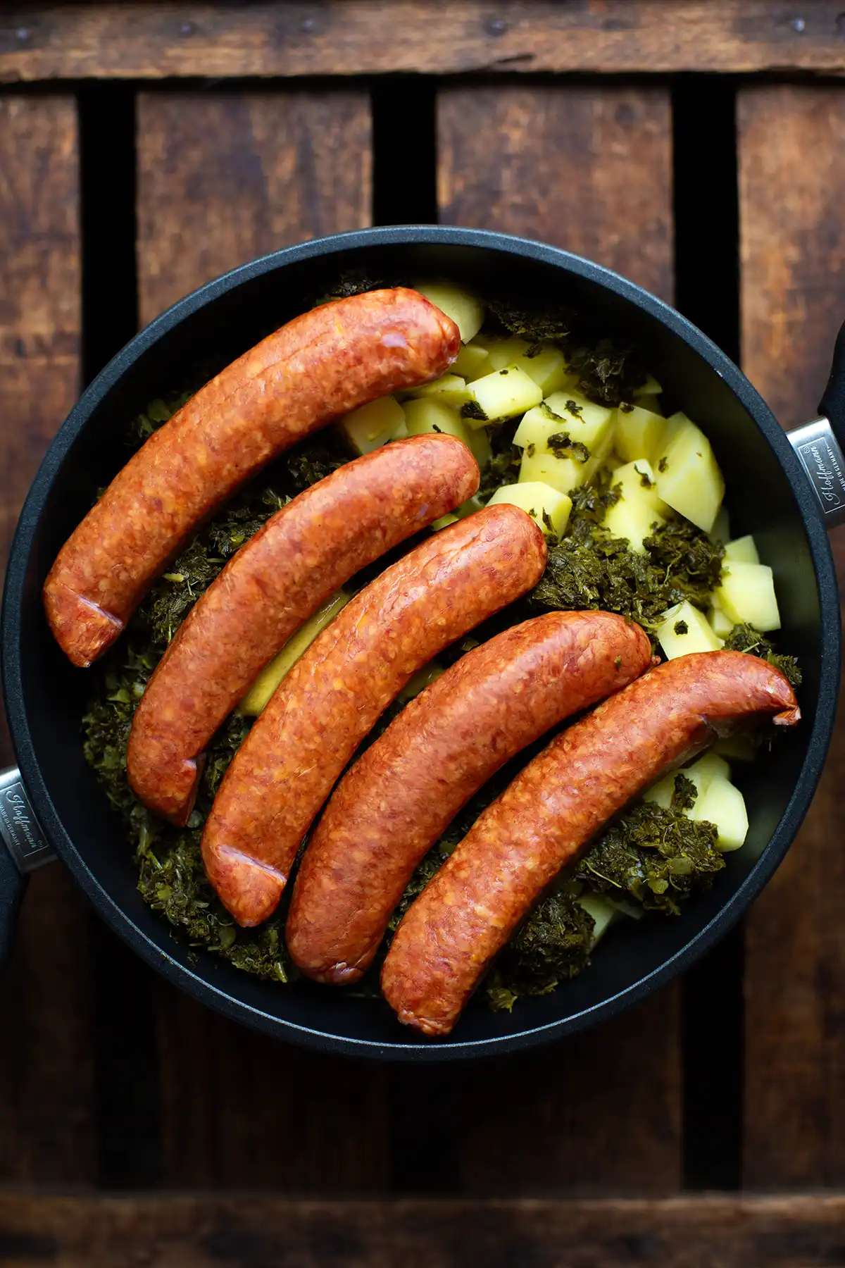 Alle Zutaten für das Grünkohl untereinander geschichtet in einem großen Schmortopf. Wir sehen Kartoffeln, vorgekochten Grünkohl, Mettenden.