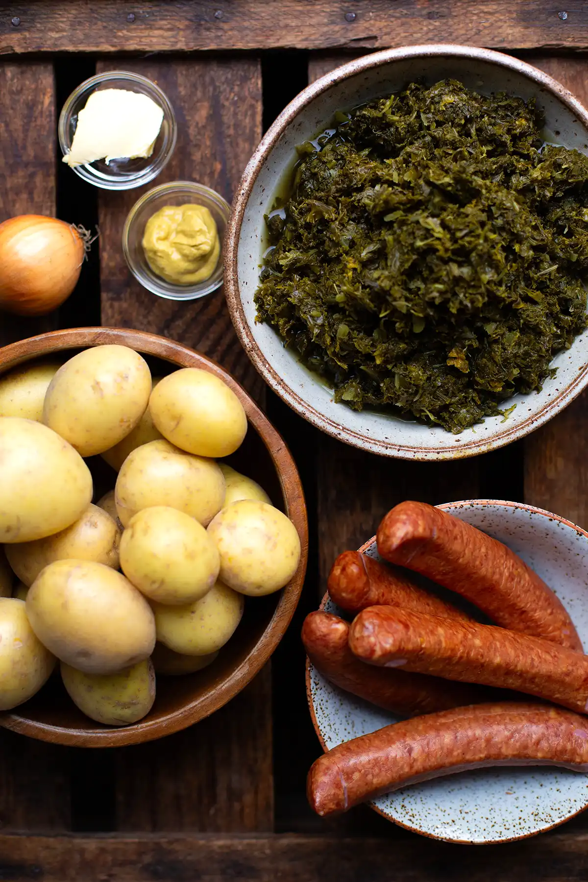 Alle Zutaten für das einfache Grünkohl Rezept auf einem Blick in kleinen Schalen auf einem braunen Untergrund. Zu sehen sind Kartoffeln, Kohlwurst, Zwiebeln, Grünkohl, Butter und Zwiebeln