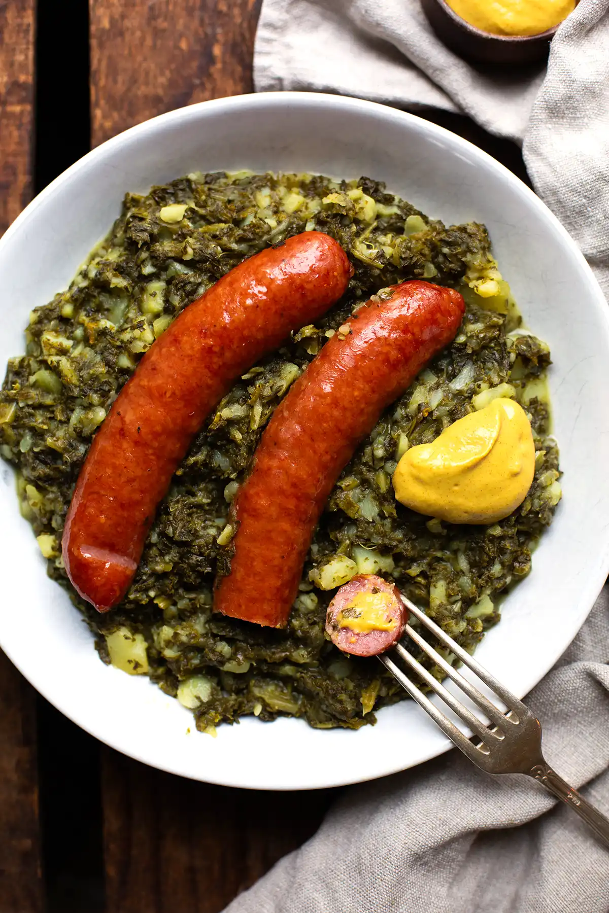 Grünkohl mit Kartoffeln, Senf und Mettenden auf einem weißen Teller fertig angerichtet. Von oben fotografiert mit angeschnittener Wurst.