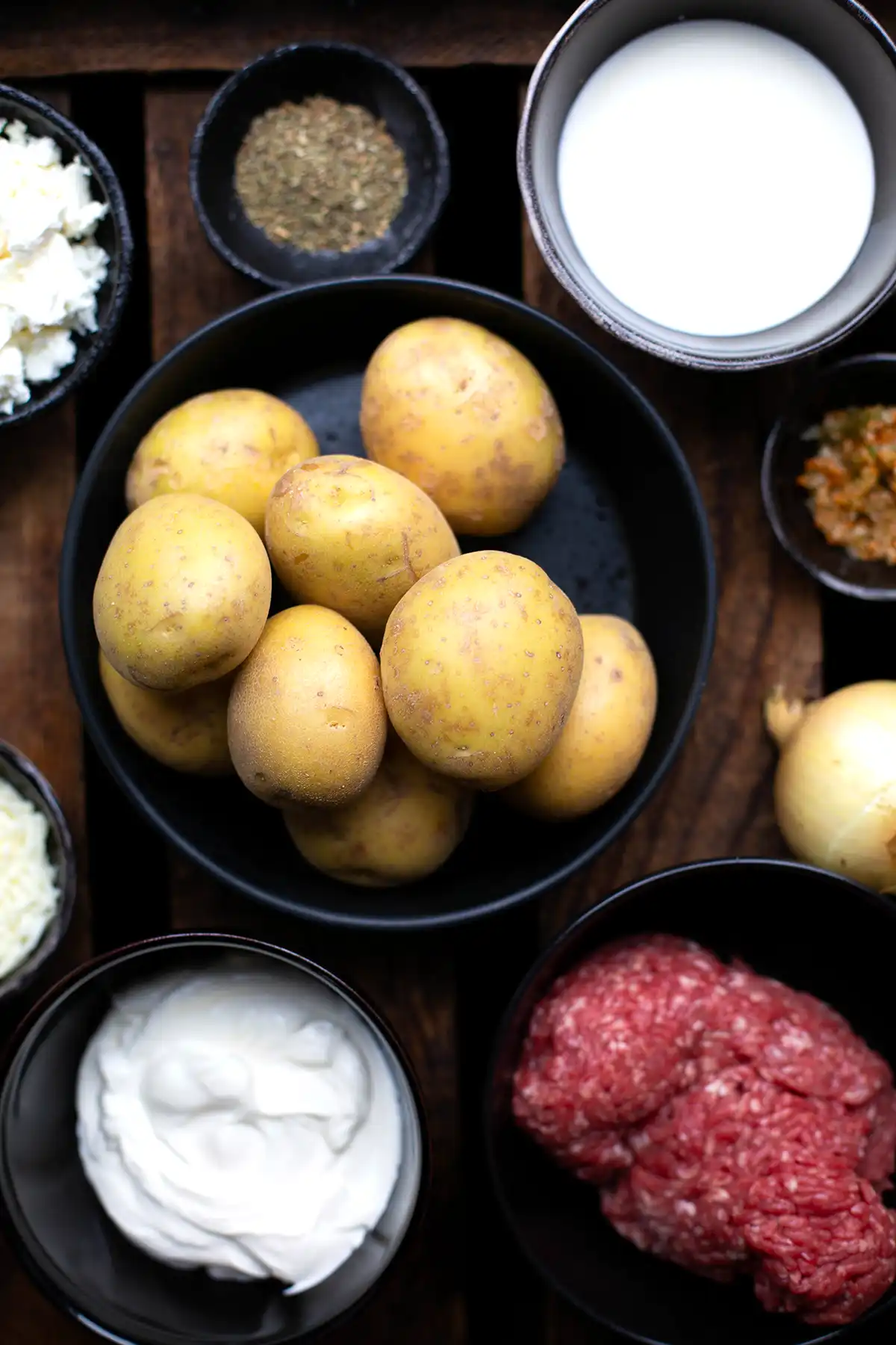 Die Zutaten für den Kartoffel-Hackfleisch-Auflauf auf einen Blick in unterschiedlichen schwarzen Schalen abgebildet. Zu sehen sind: Kartoffeln, Zwiebeln, Schafskäse, Sahne, Gemüsebrühpaste, Oregano, Hackfleisch, Mozzarella und Schmand.