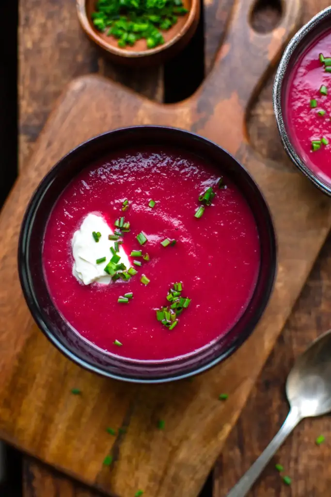 Fertige, pürierte Rote-Bete-Suppe in einer dunklen Schüssel, garniert mit Schmand und Schnittlauch auf einem rustikalen Holzbrett