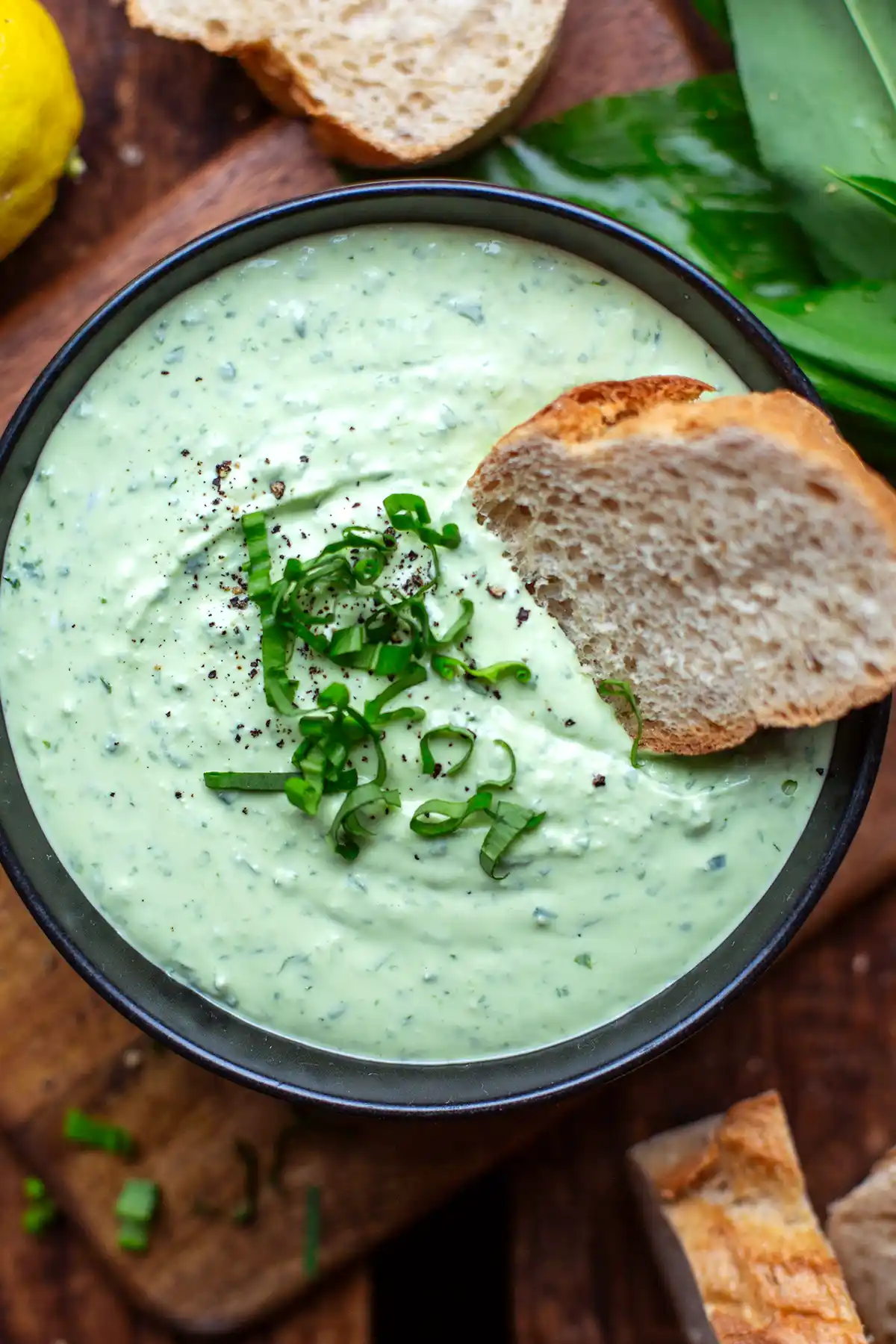 Eine schwarze Schüssel mit cremigem Bärlauchdip, dekoriert mit frischen Bärlauchblättern auf einem Holztisch.