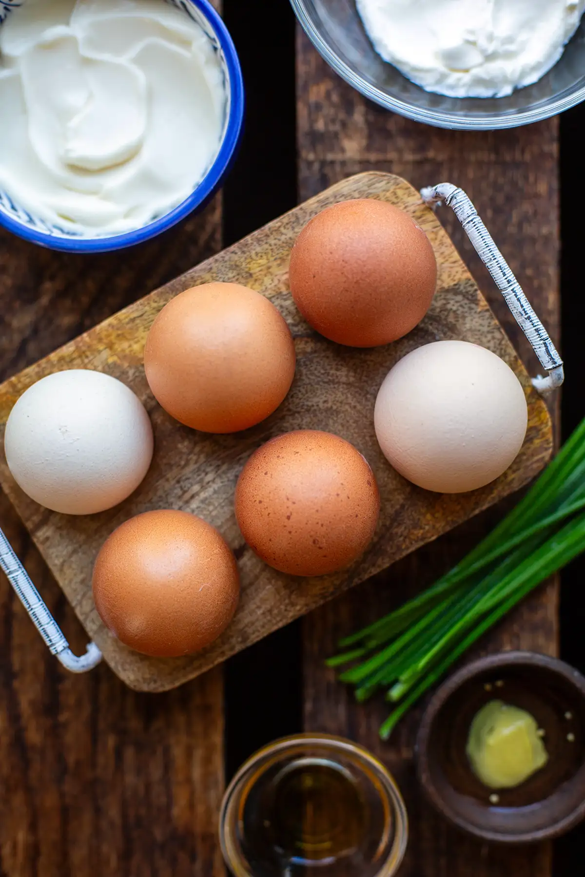 Zutaten für den einfachen Eiersalat auf einem dunklen Holzuntergrund. Zu sehen sind 6 hartgekochte Eier, Ahornsirup, Senf, Schnittlauch, Schmand und Mayonnaise.