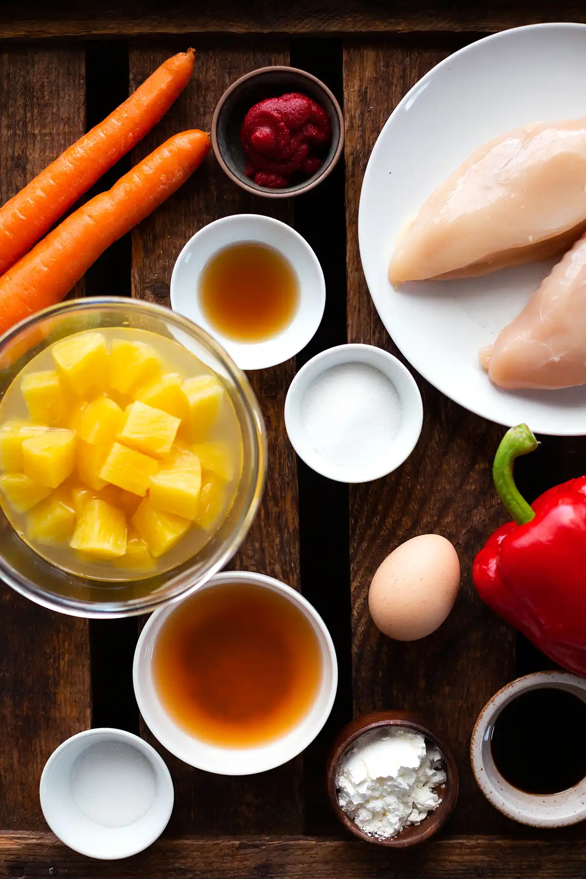 Die Zutaten für Hähnchen süß-sauer: Karotten, Ananas, Tomatenmark, Hähnchen, Speisestärke, Ei, Paprika, Essig, Sojasoße, Sesamöl und Salz, teilweise in Schälchen.