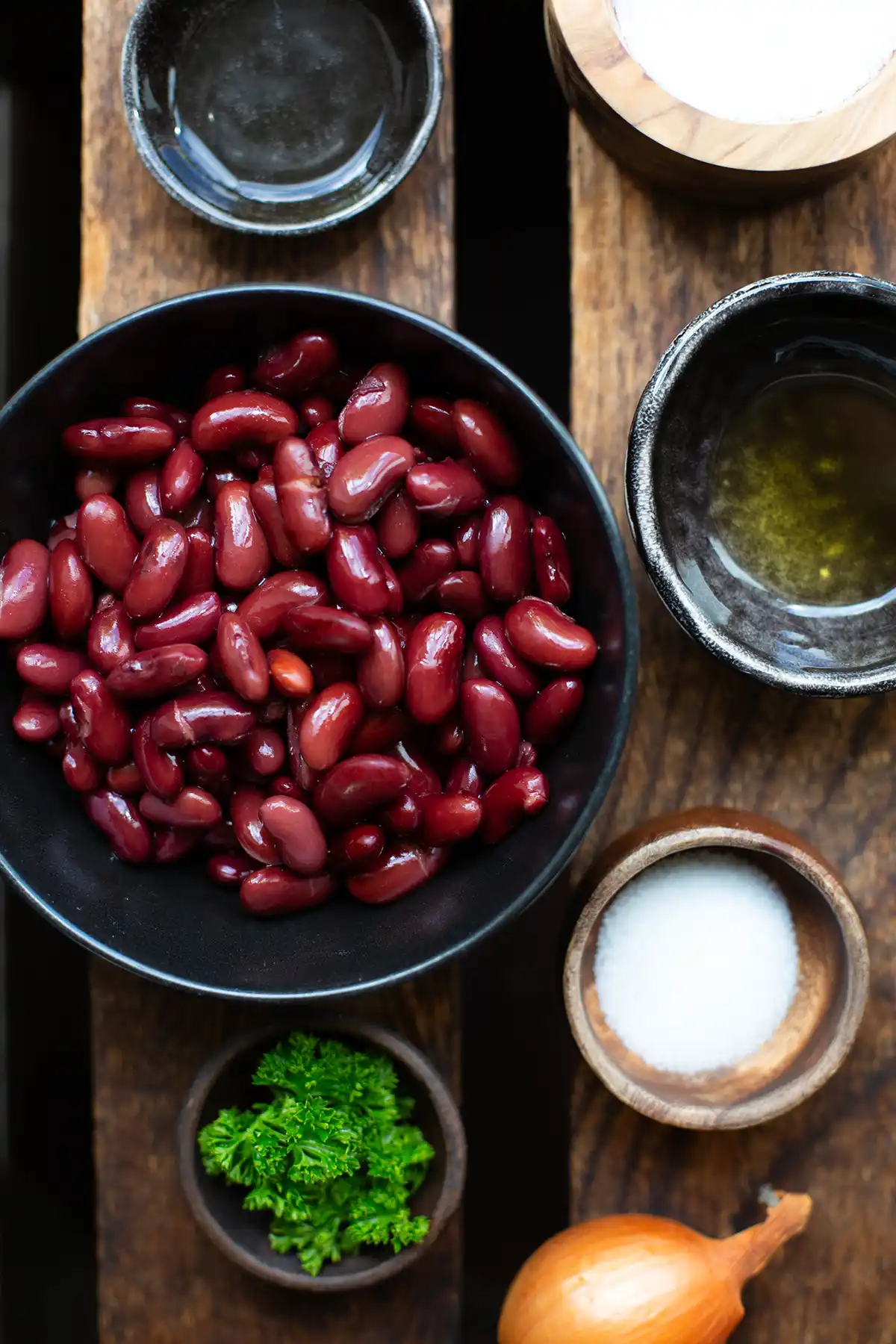 Alle Zutaten für den Kidneybohnensalat einzeln in Schüsseln angerichtet: Kidneybohnen aus der Dose, eine Zwiebel, Walnussöl, dunkler Balsamico, Salz, Pfeffer und Petersilie als Deko