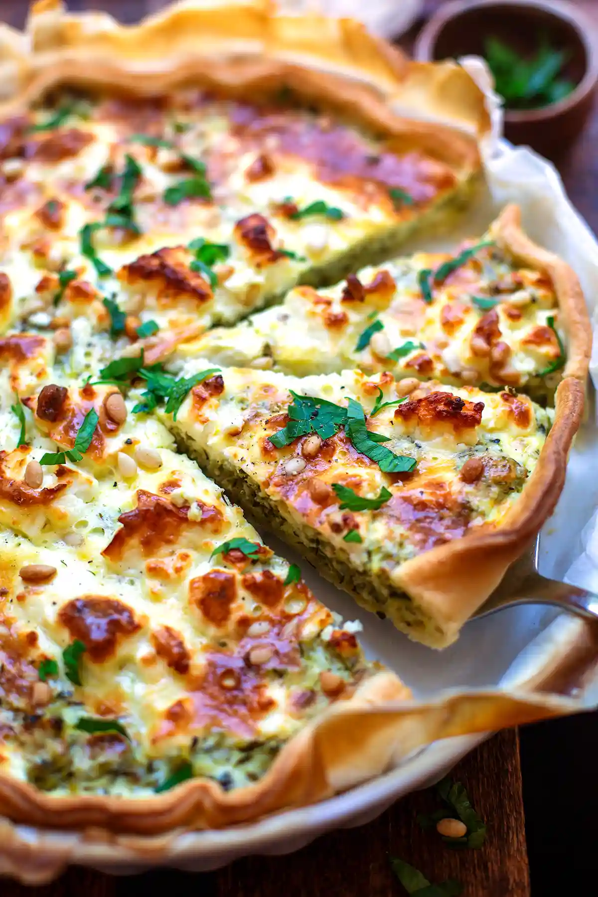 Angeschnittene Zucchini-Quiche mit Feta und Pinienkernen in der Quicheform, ein Stück wird angehoben