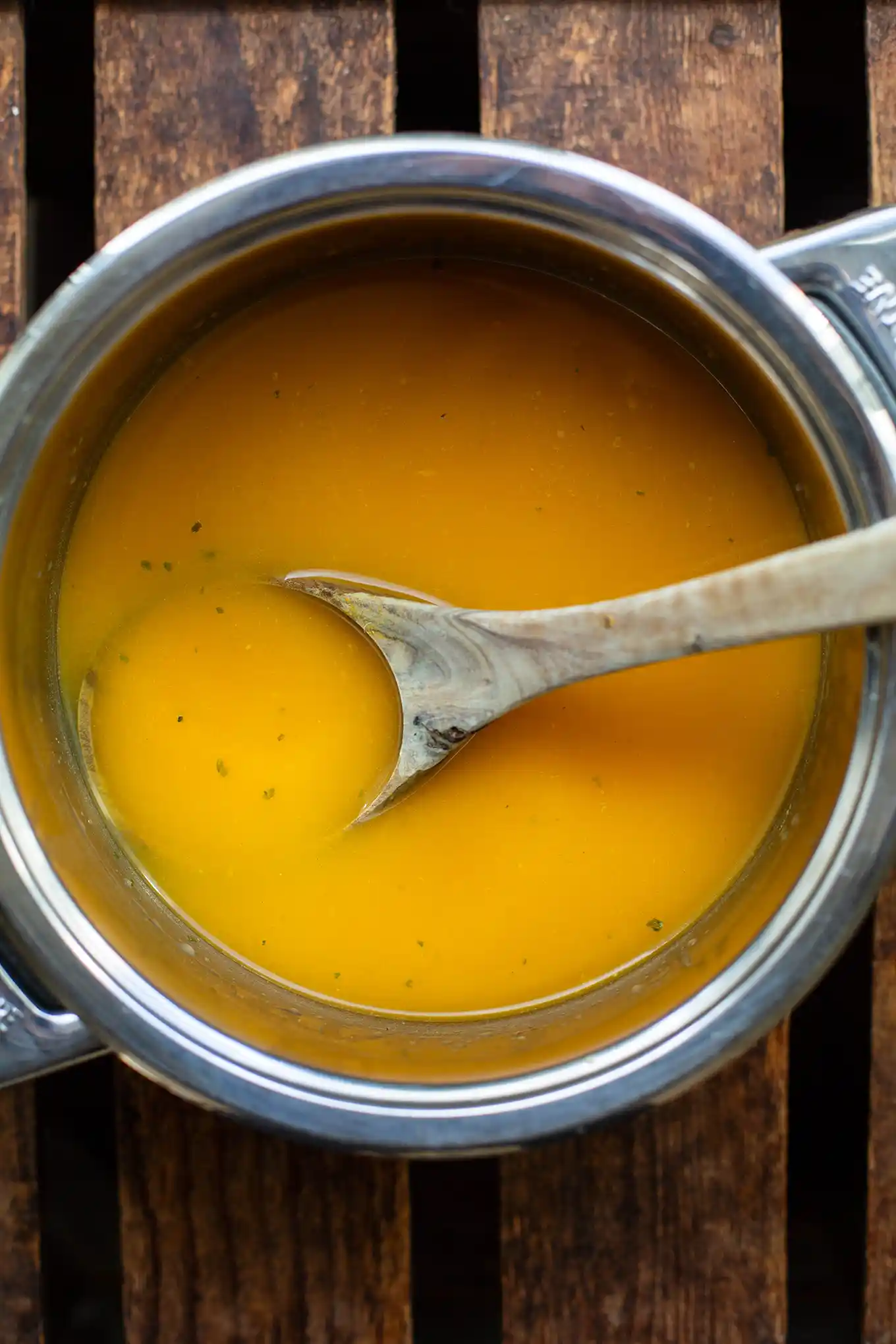 Püriertes Gemüse mit Gemüsebrühe in einem silbernen Topf auf einem Holztisch und mit einem Holzlöffel in der Suppe. Man sieht kleine Kräuter